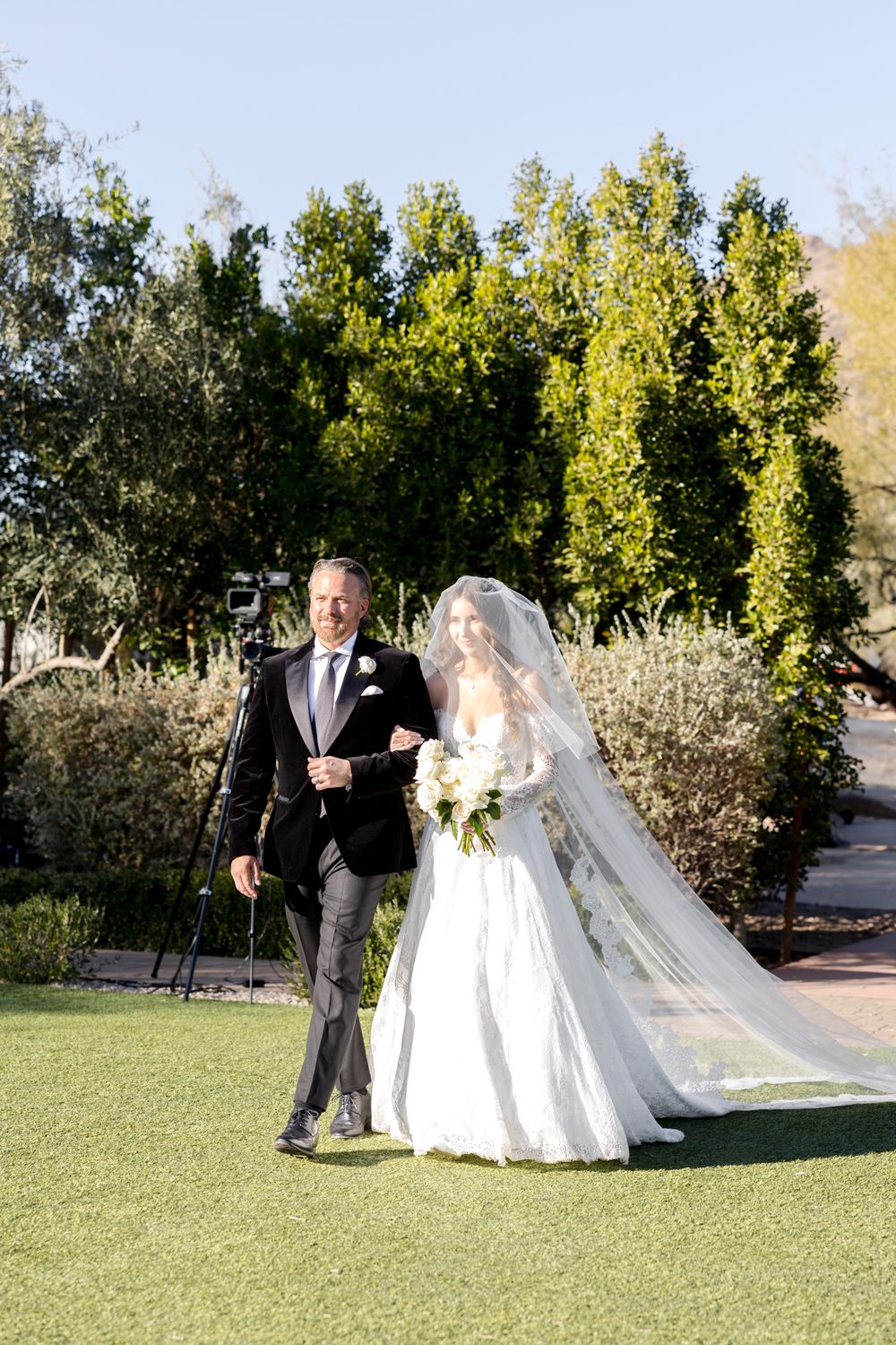 A couple in formal wedding attire walking together on a grassy lawn with trees in the background.