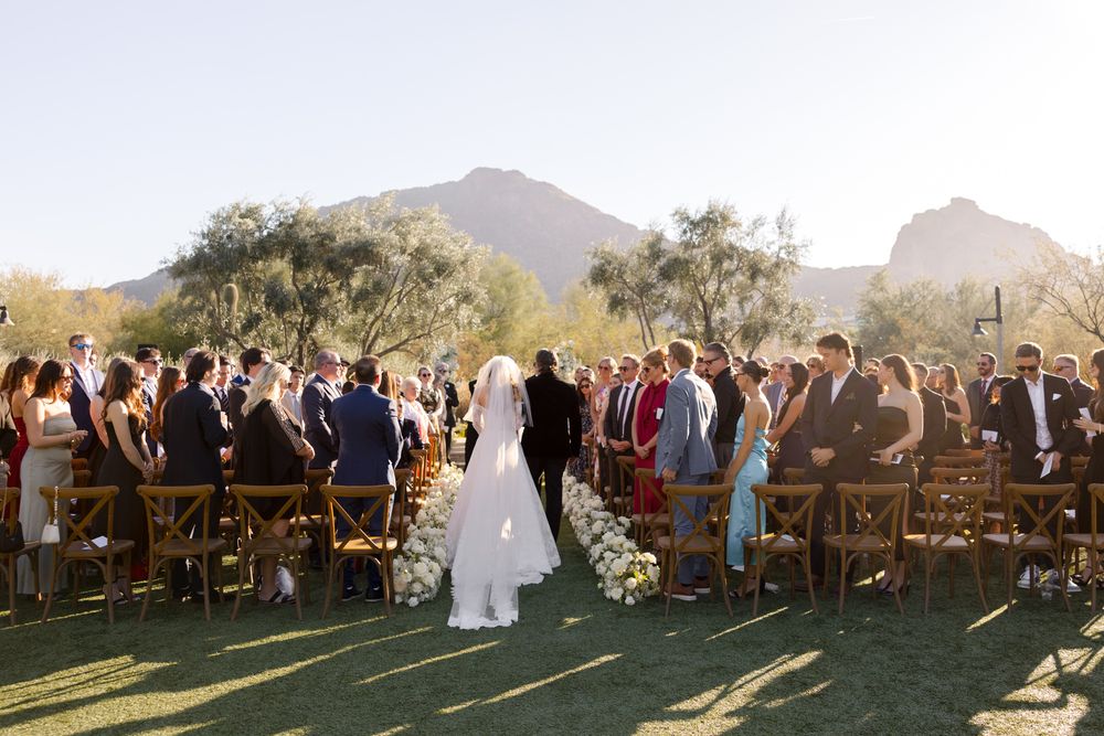 Wedding ceremony guests stand in a semicircle on a grassy lawn with mountain peaks and golden desert landscape in background.