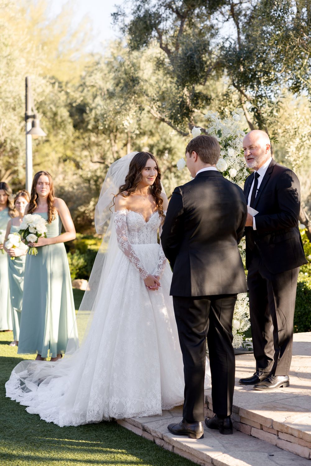 An outdoor wedding ceremony takes place in a garden setting with a bride in a long-sleeved lace gown and attendants in mint green.