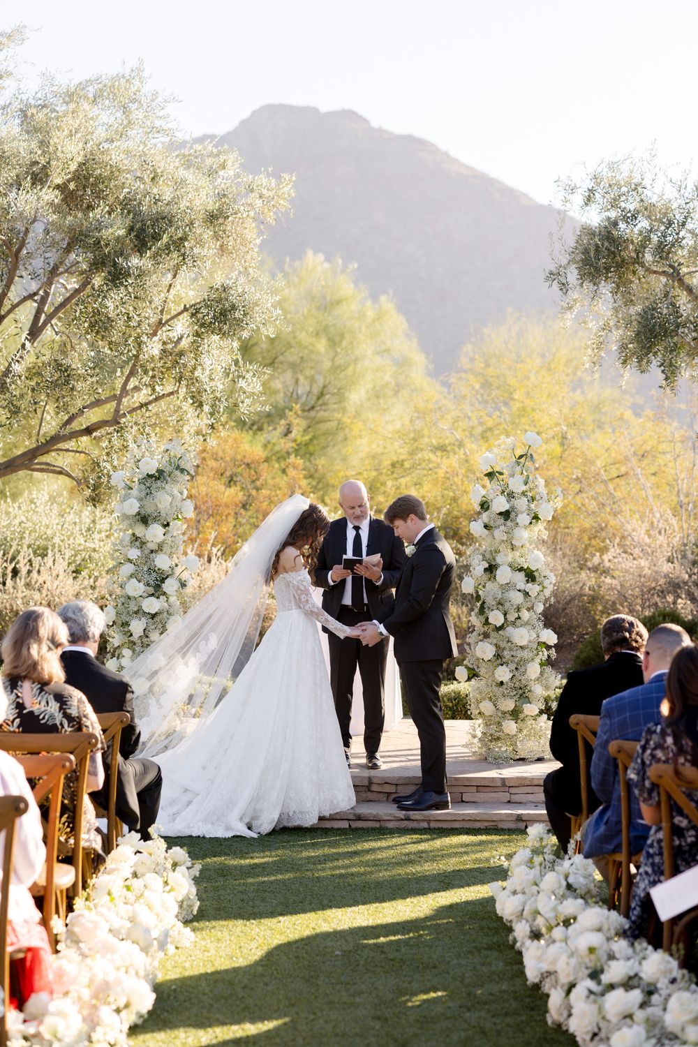 An outdoor wedding ceremony with lush floral arrangements takes place against a scenic mountain backdrop in golden afternoon light.