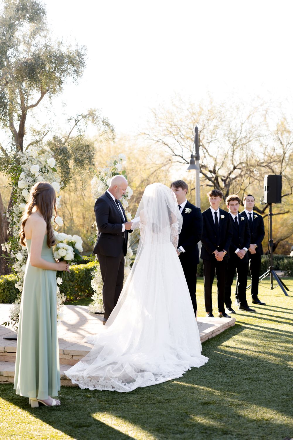 Wedding ceremony on a sunlit lawn with attendants in mint green and black formal attire.