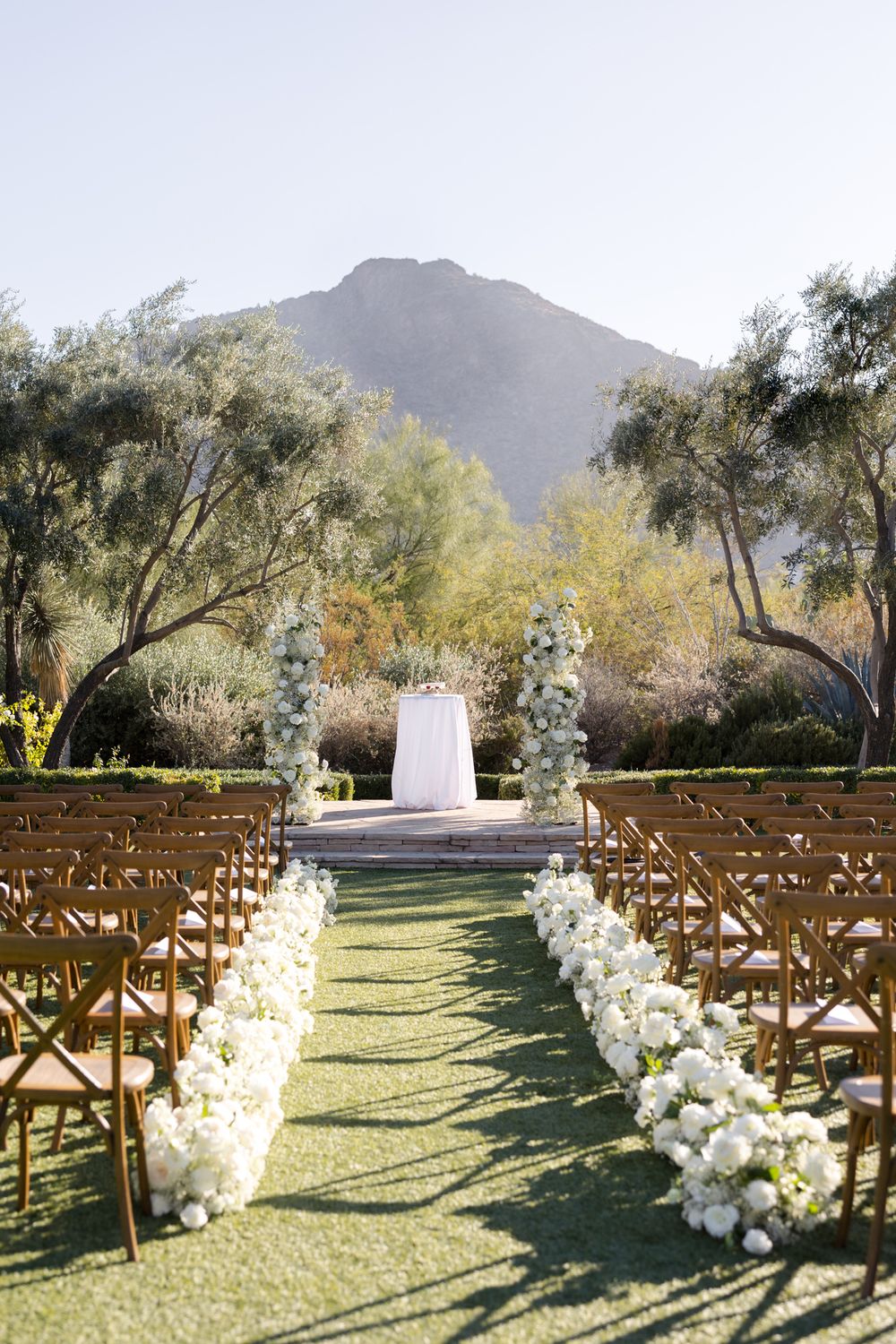 Outdoor wedding ceremony setup with wooden chairs and white floral aisle arrangements against a mountain backdrop.