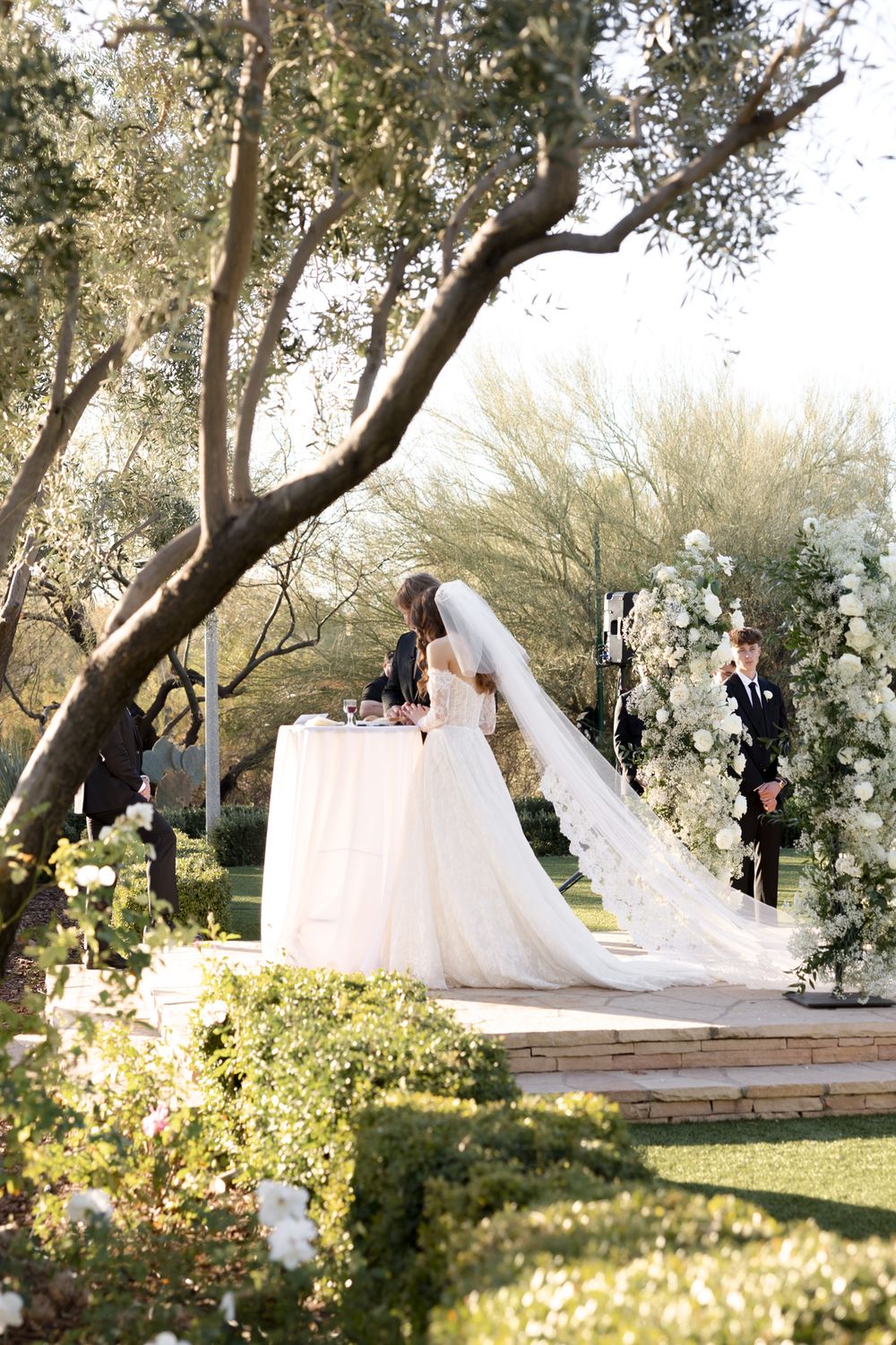 A wedding ceremony takes place in a garden setting with a floral arch and long flowing veil against a sunlit backdrop.