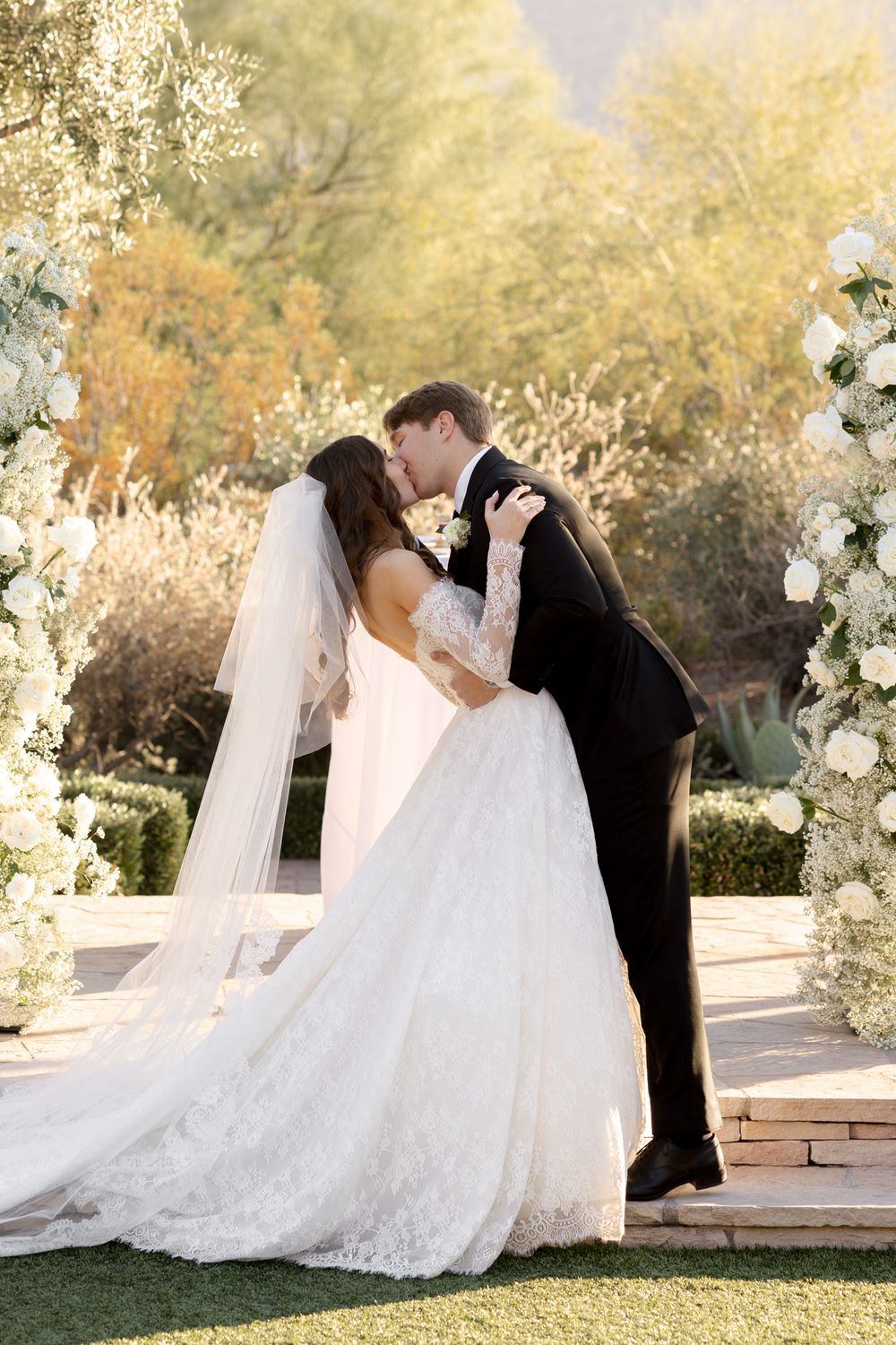 A romantic fall wedding scene with couple kissing under a floral arch decorated with white roses in golden sunlight.