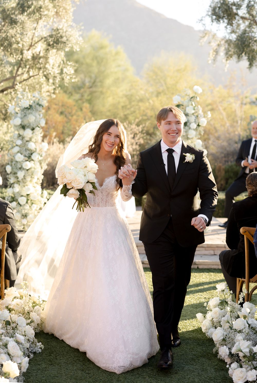 Newlyweds walk down the aisle at outdoor garden wedding with white floral decorations and mountain backdrop.
