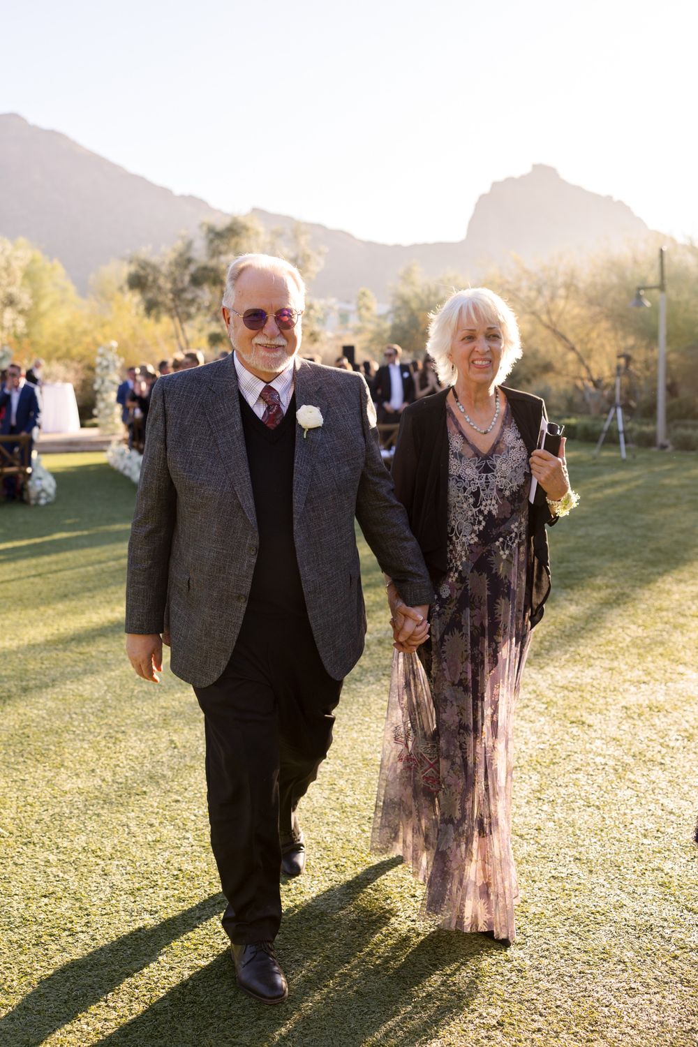 An elderly couple poses together on a sunny outdoor lawn with mountains in the background during a wedding celebration.