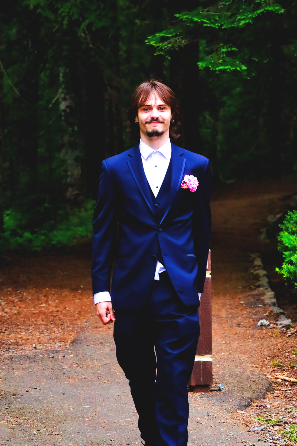 Person wearing navy blue suit with pink boutonniere stands on forest path.