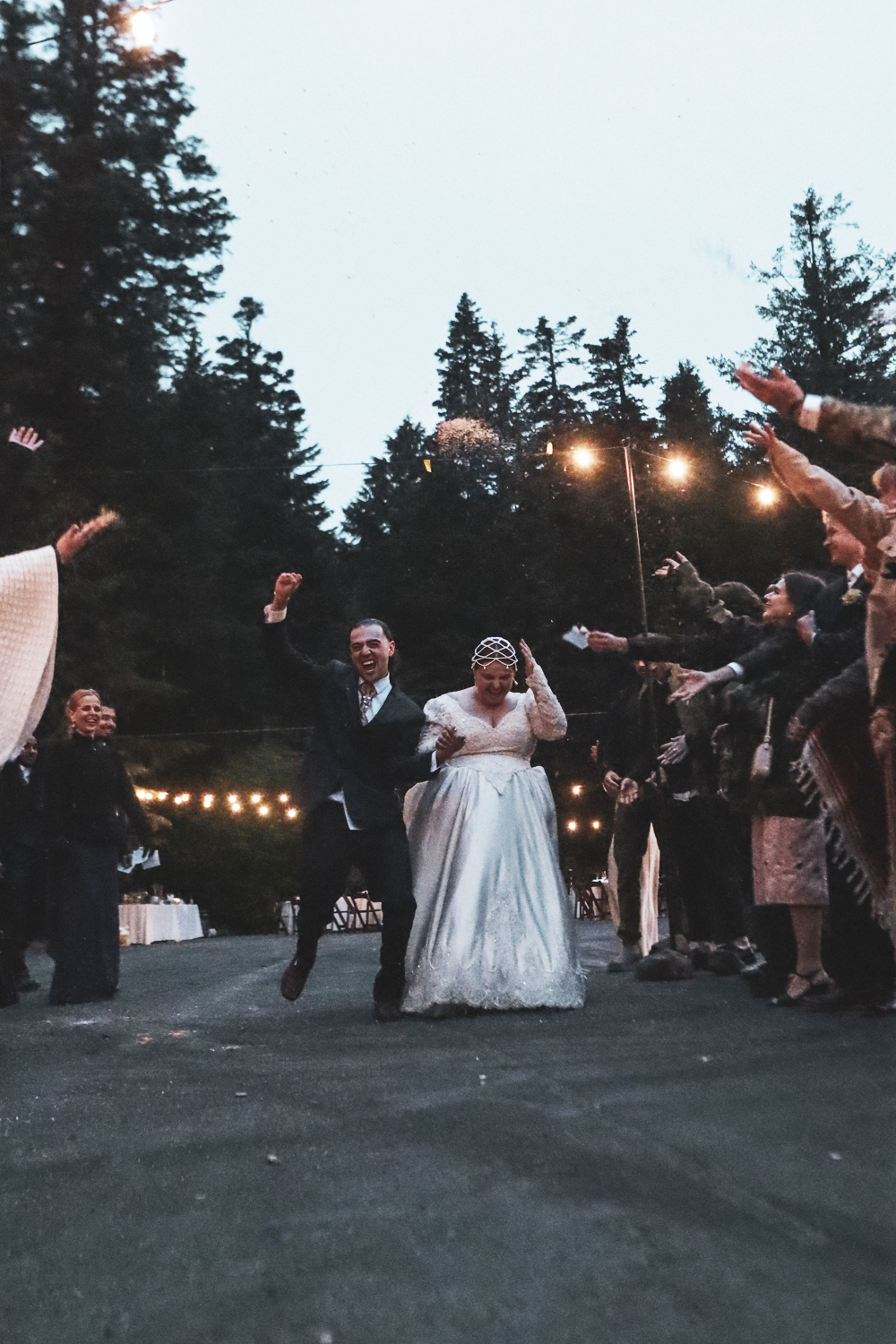 Newlywed couple walking through sparkler exit at outdoor evening wedding reception with pine trees in background.