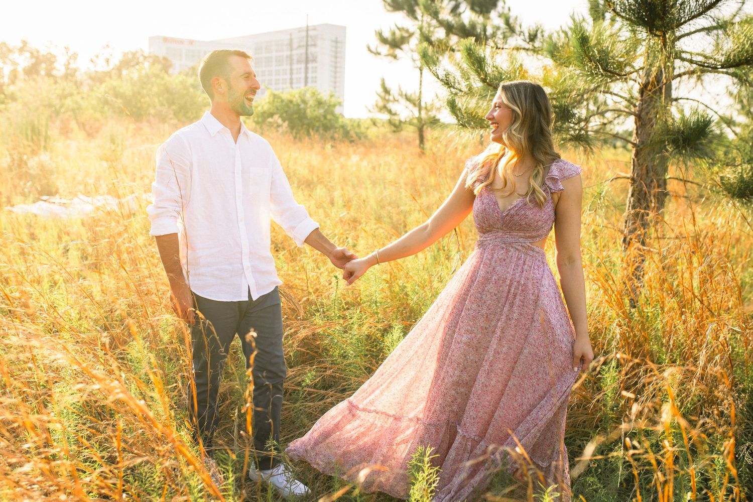 Couple shares romantic moments in golden sunlit meadow wearing pink dress and white shirt during sunset photoshoot.