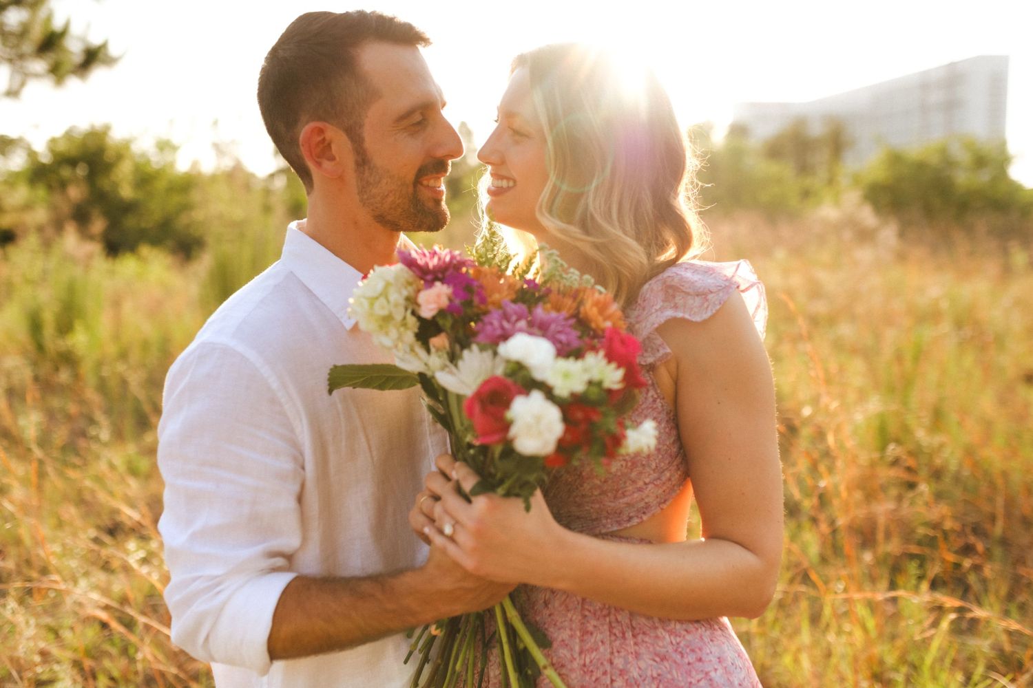 A couple embraces in a sunlit field surrounded by golden grass and wildflowers at sunset.