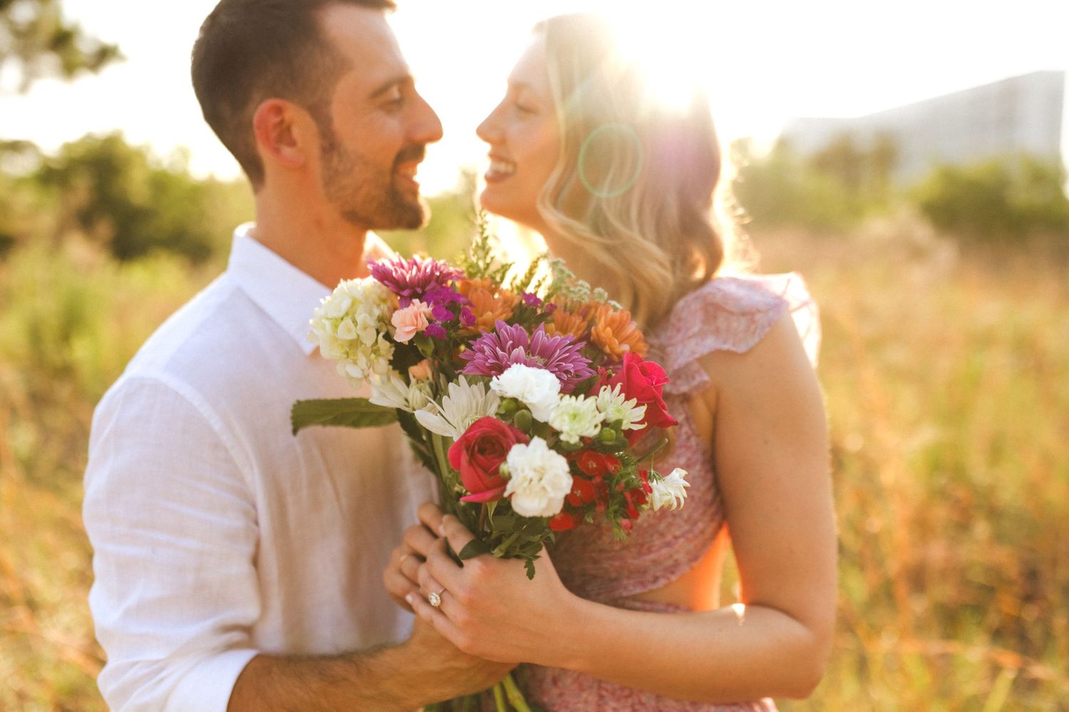 Couple laughing and embracing in golden sunlit field while holding colorful wildflower bouquet at sunset.