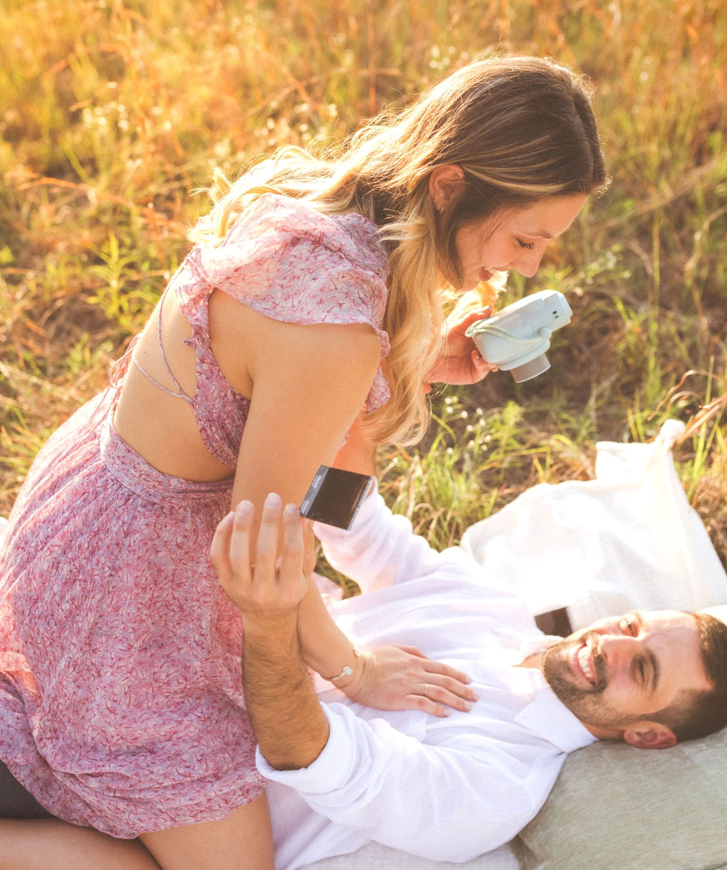 A couple in a romantic moment outdoors, with someone in a pink floral dress leaning over their partner lying on white fabric.