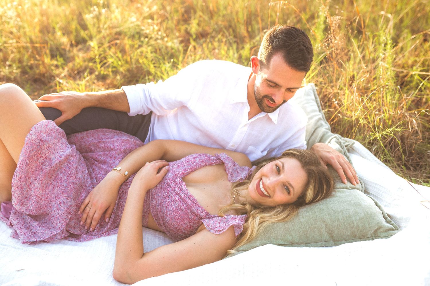 A couple relaxes together on a pillow in a sunlit field of tall grass during sunset.