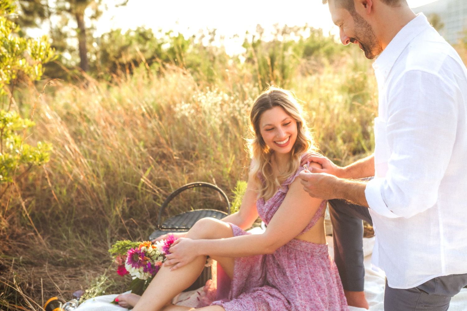 A couple sits together at sunset in a golden field with a picnic basket and flowers on a warm summer evening.