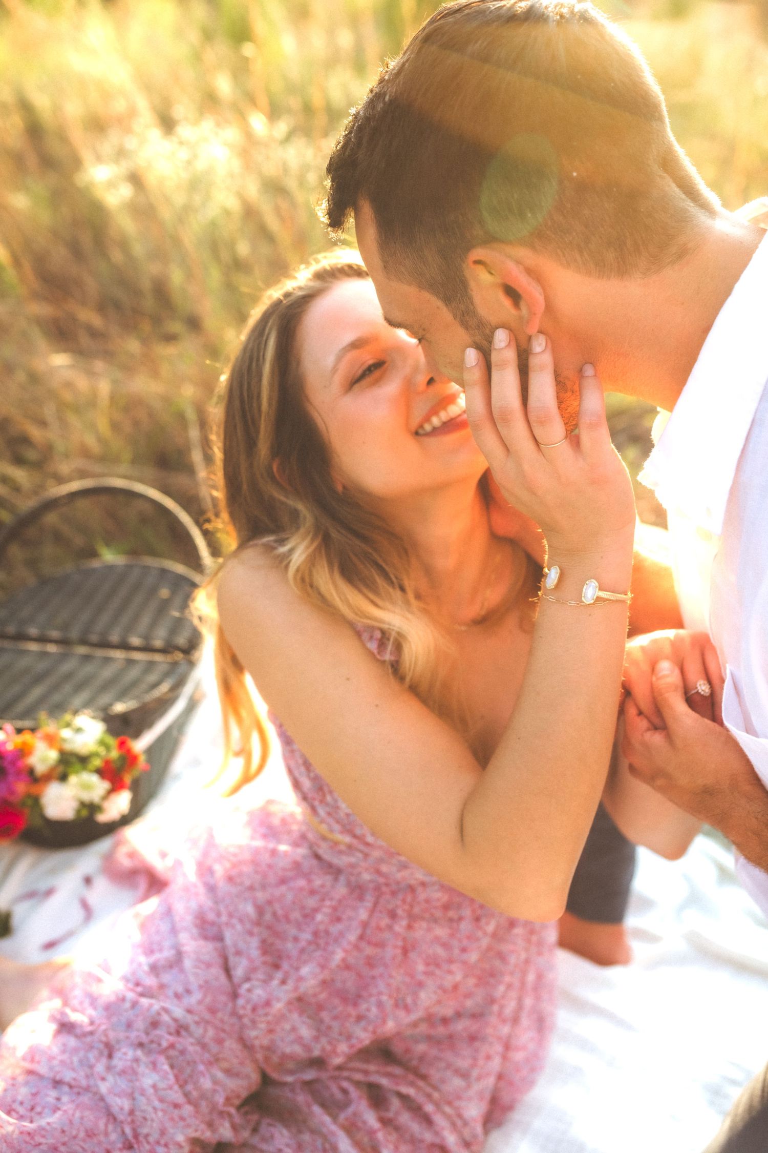 Romantic couple sharing tender moment at sunset picnic with flowers and wicker basket on blanket.