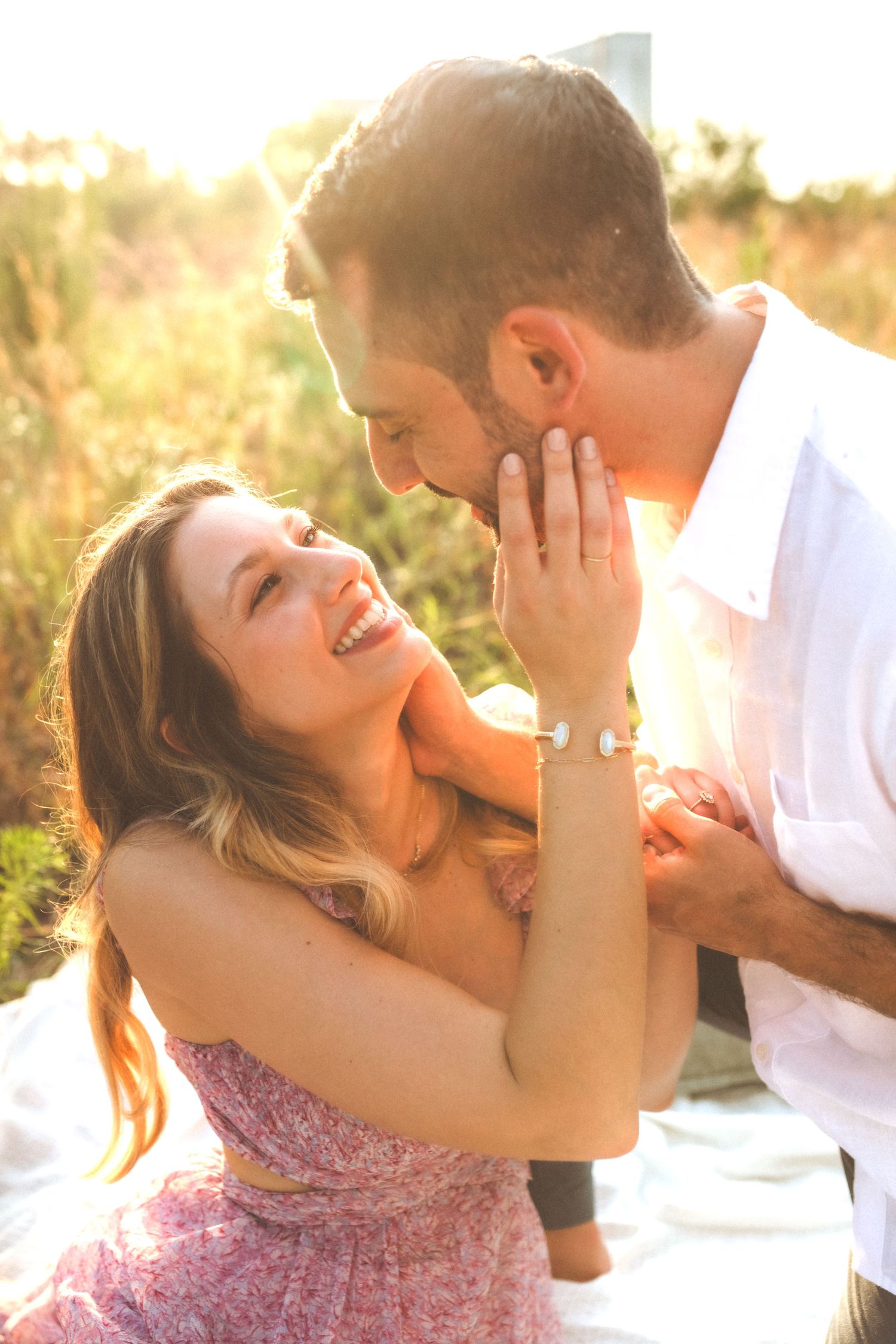 A couple shares a tender romantic moment at golden hour, laughing together in a sun-drenched field.