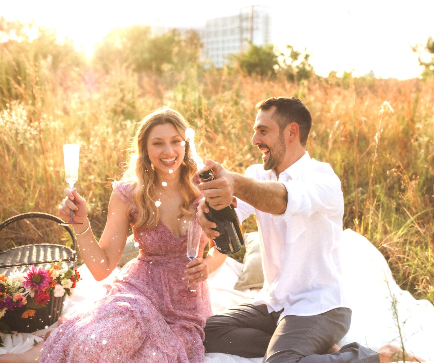 A couple enjoys champagne during a romantic sunset picnic in a golden meadow with a floral arrangement nearby.