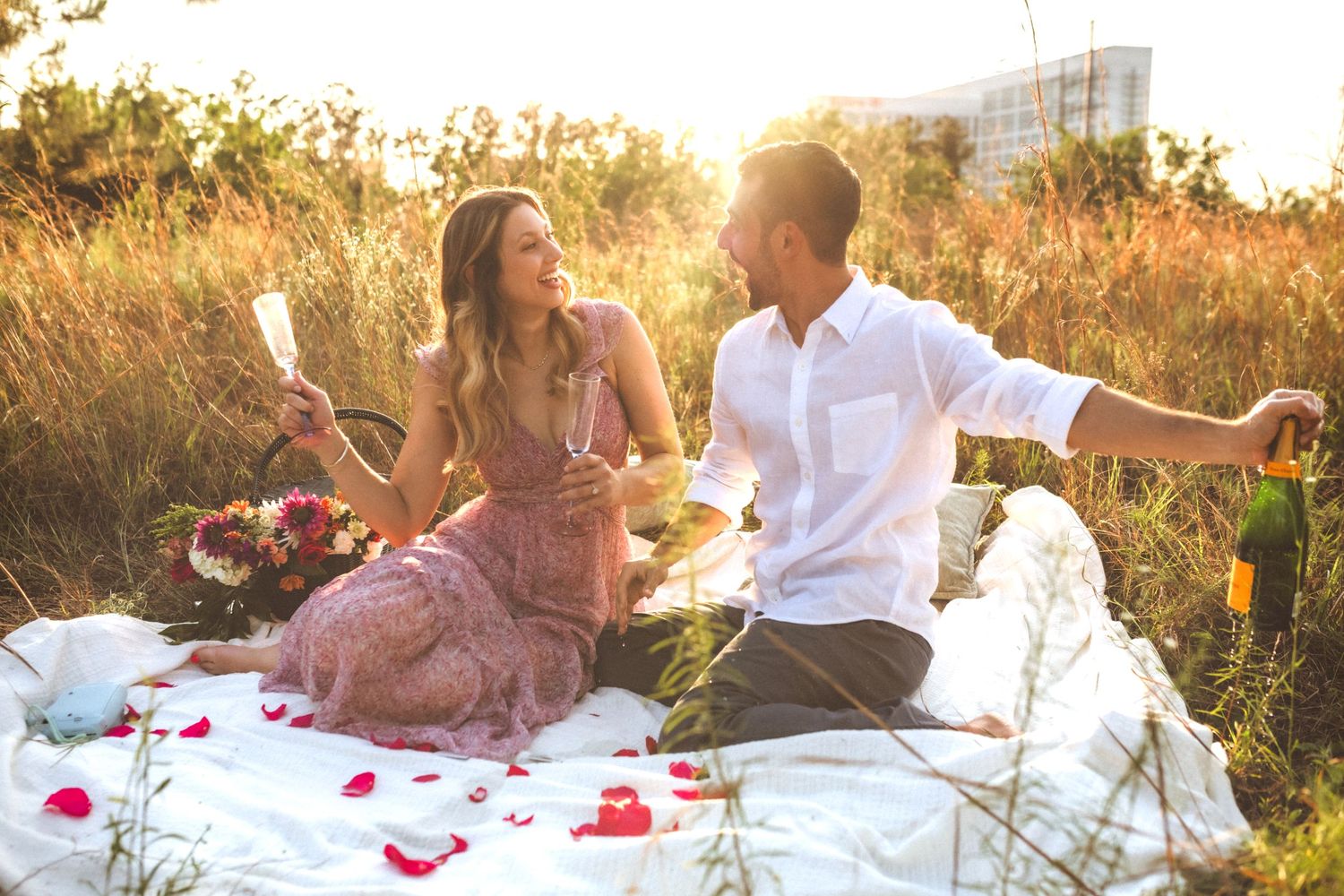 Couple having romantic sunset picnic on blanket with rose petals scattered around in golden field.
