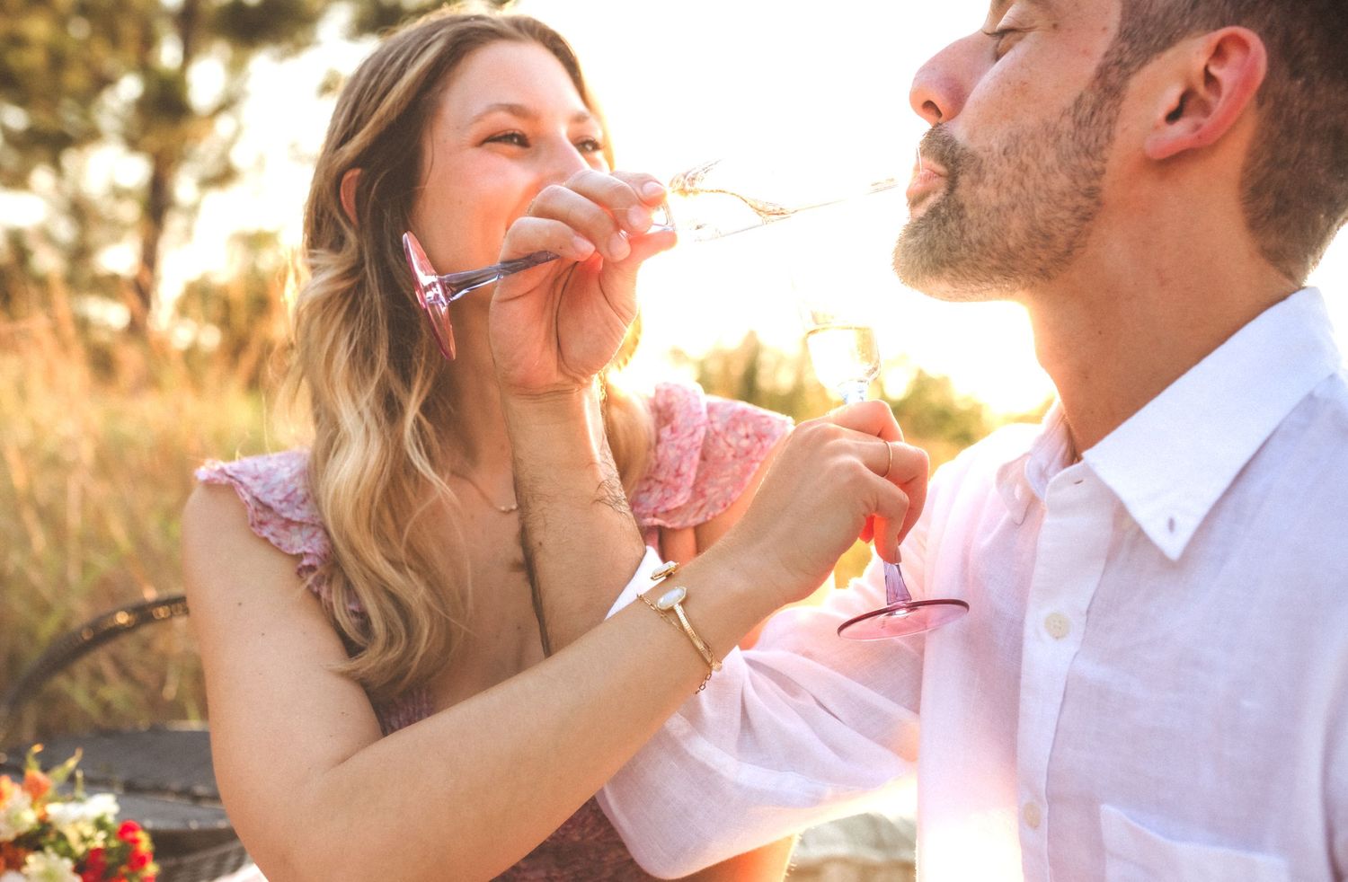 Romantic couple sharing drinks at sunset in golden meadow during intimate picnic moment.