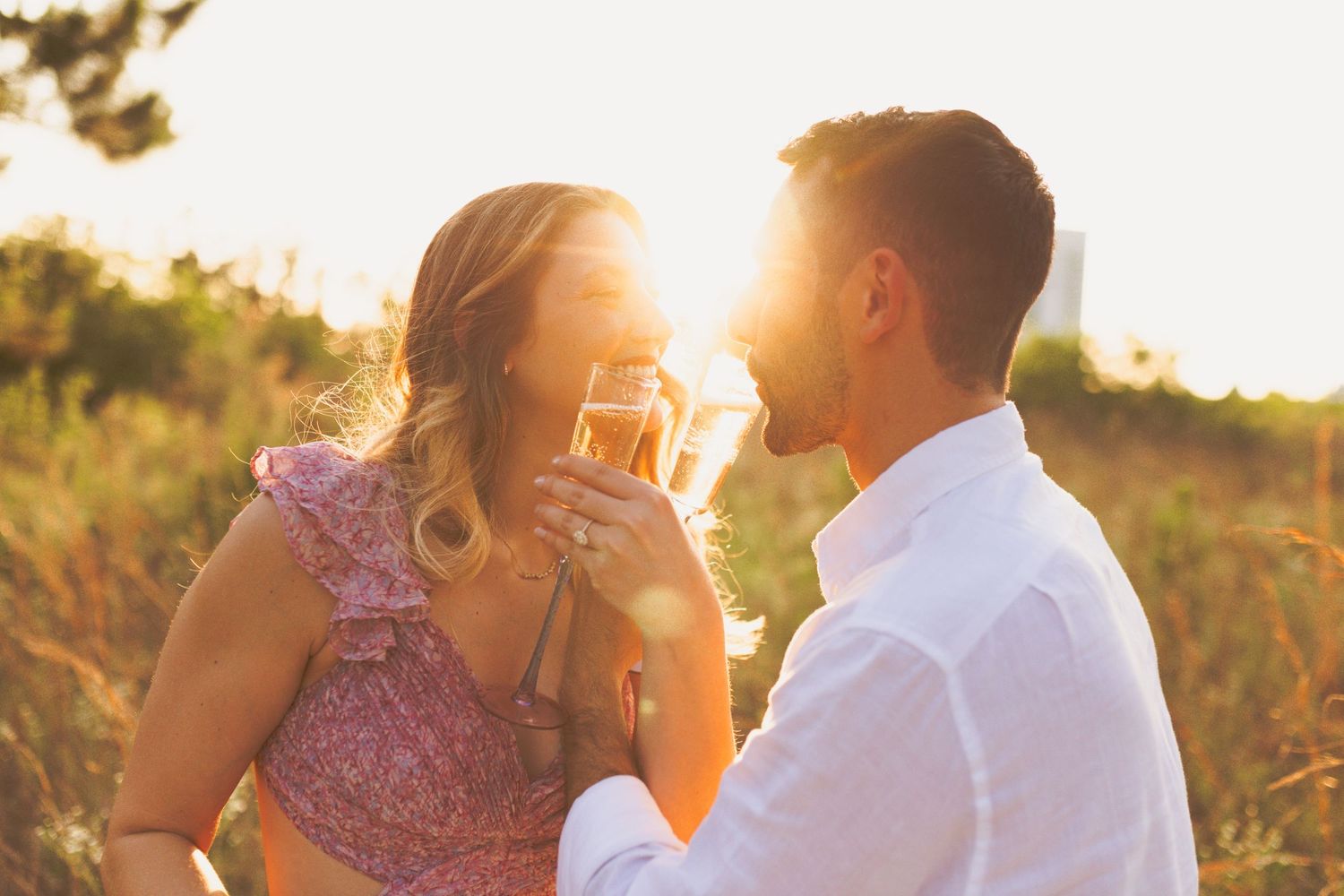 A couple in flowy pastel clothing embraces in a golden sunlit meadow during sunset.