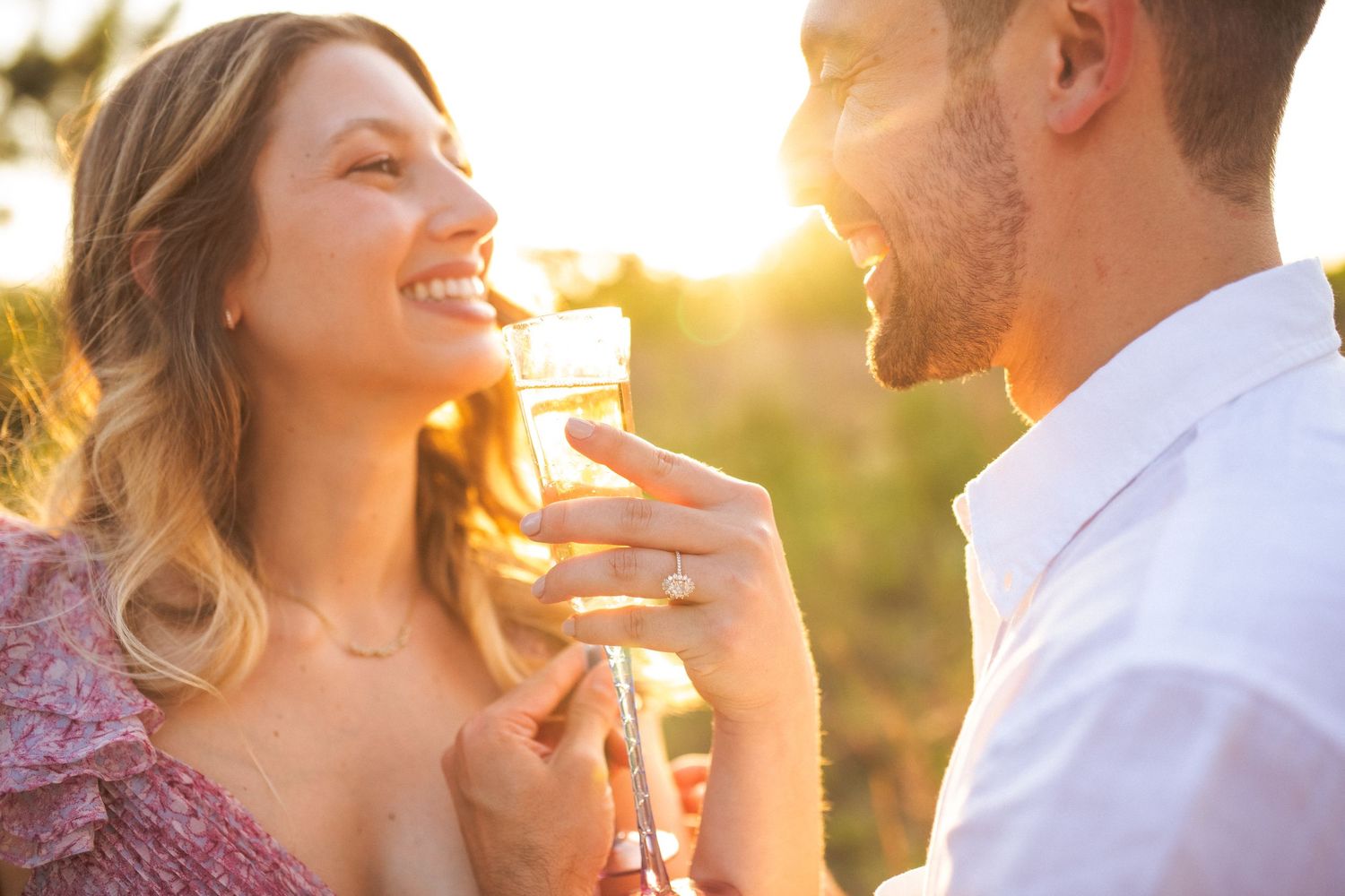 A happy couple shares a romantic moment in a sunlit field at golden hour, both smiling and enjoying wine together.