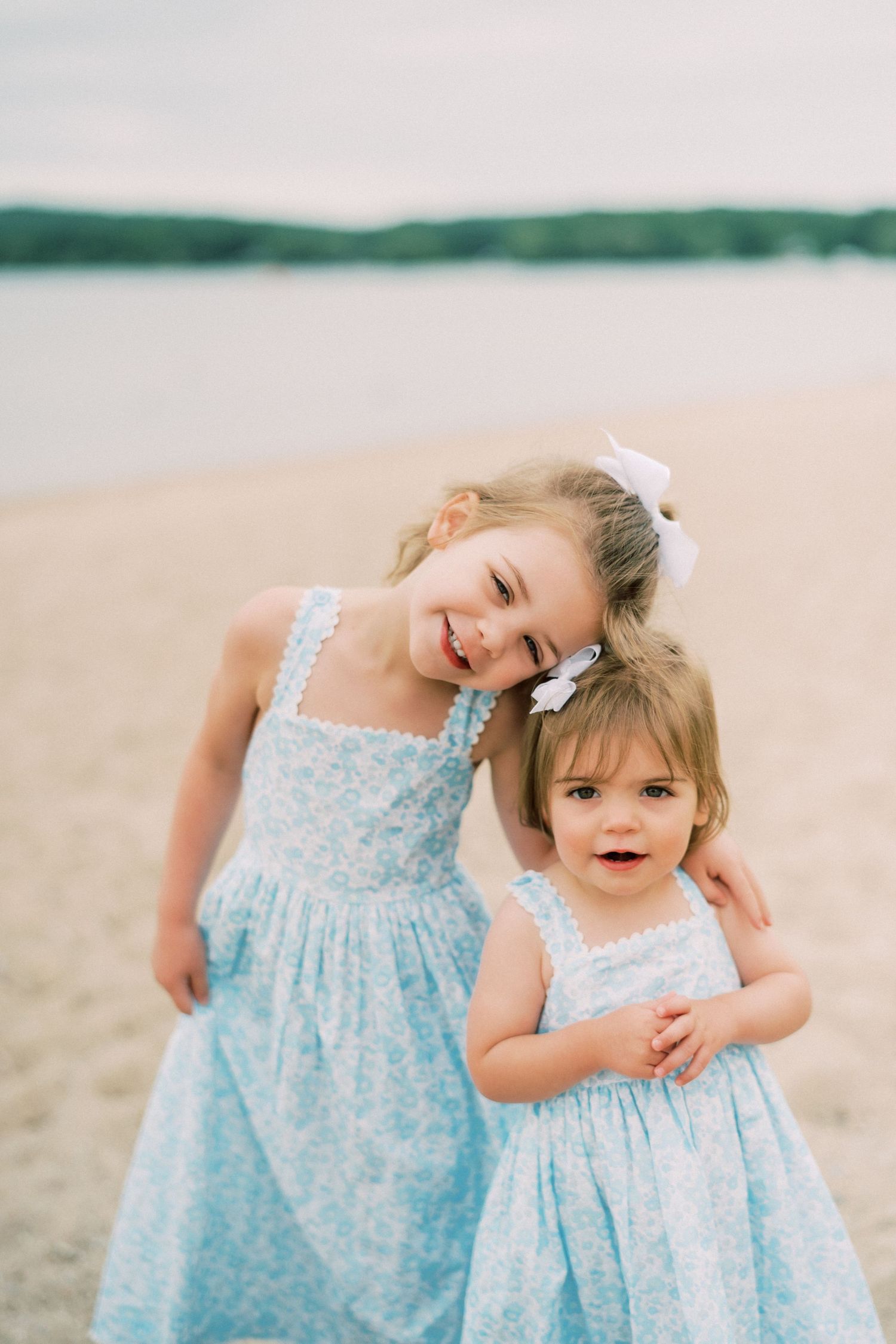 Two little girls in matching light blue floral dresses pose together on a beach with a lake in the background.