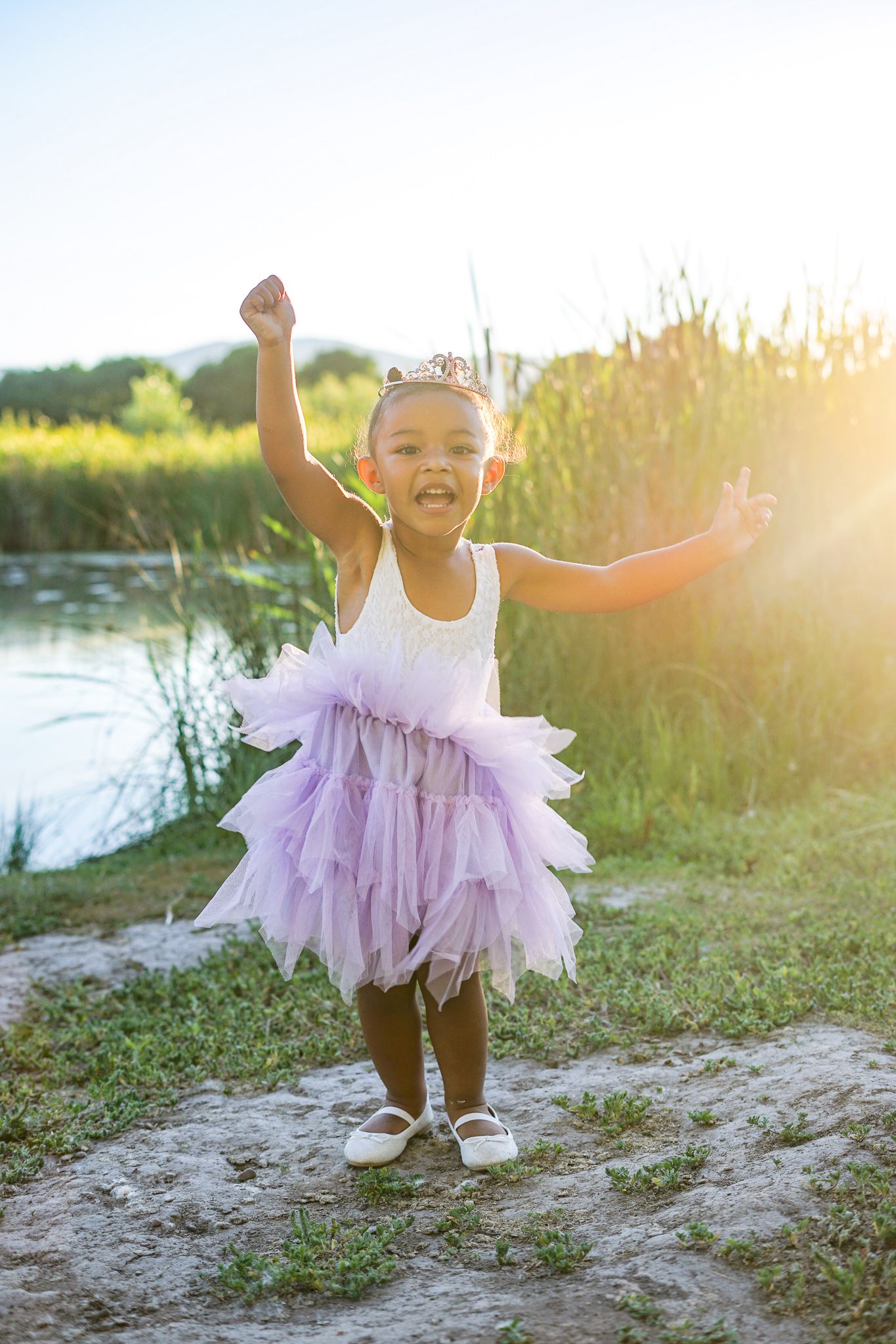 Young dancer in purple tutu dress poses joyfully by lake at sunset.