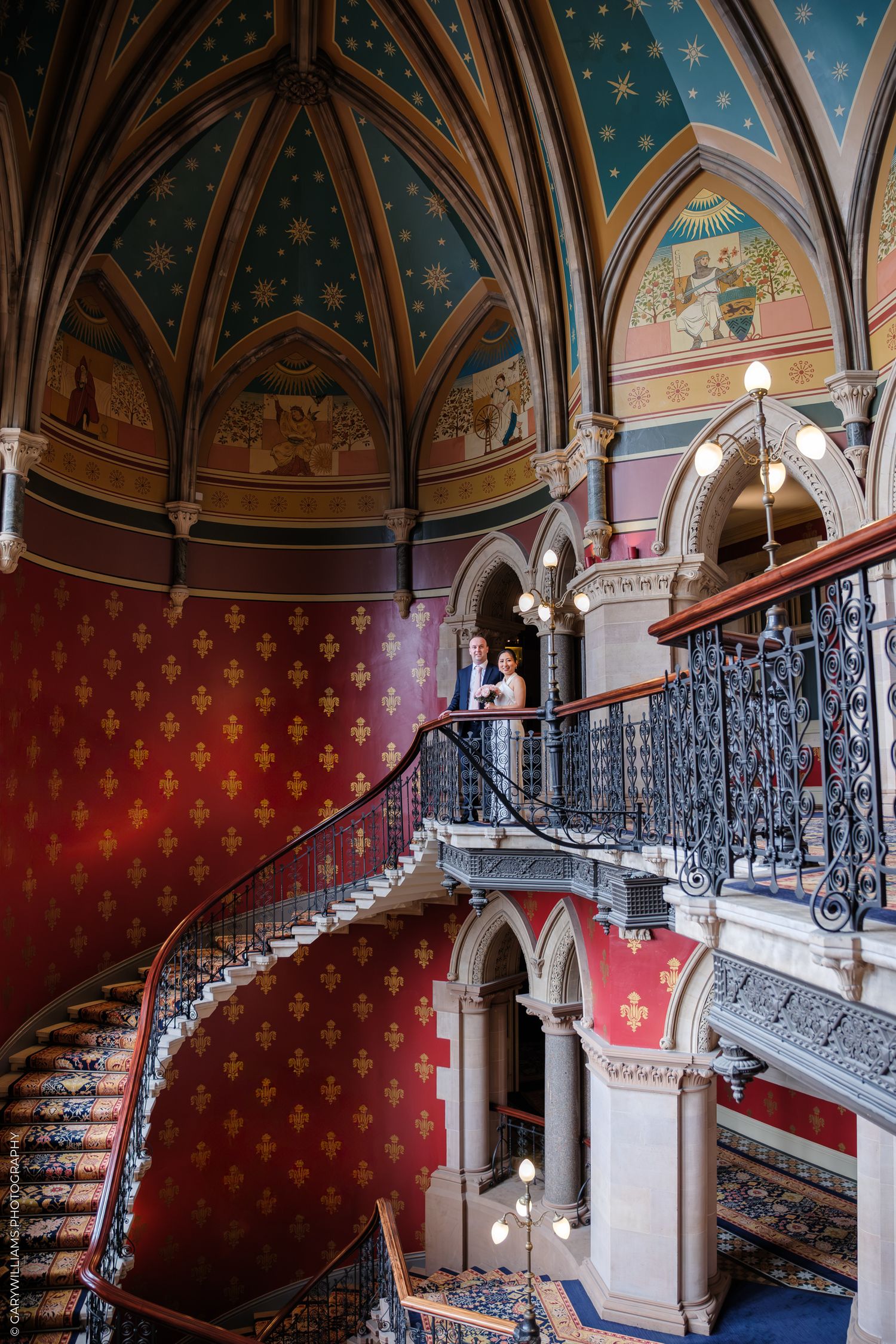 Ornate Gothic staircase with red carpeting and blue ceiling in historic St. Pancras Hotel features detailed iron railings.