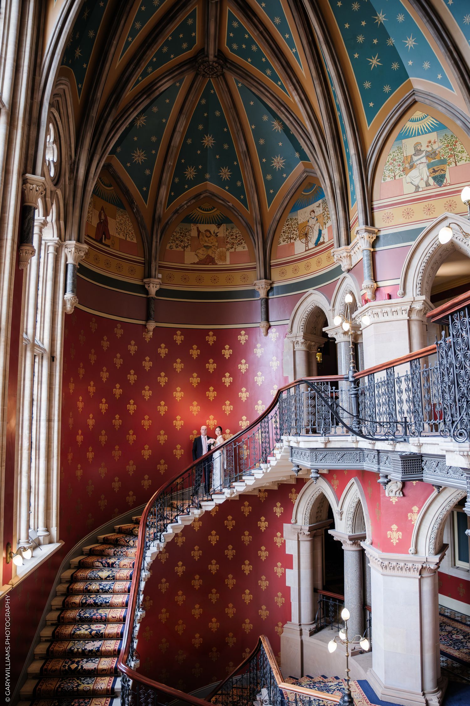 Ornate Gothic staircase with red fleur-de-lis wallpaper and blue starry ceiling in historic mansion interior.