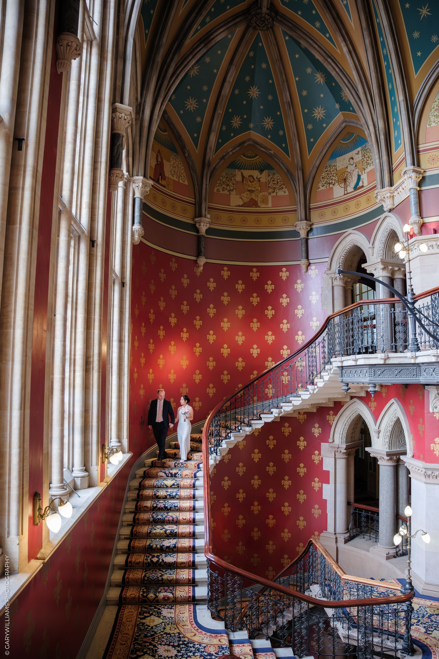 Elegant carpeted staircase with red fleur-de-lis wallpaper inside a Gothic Revival castle featuring vaulted ceilings and arches.