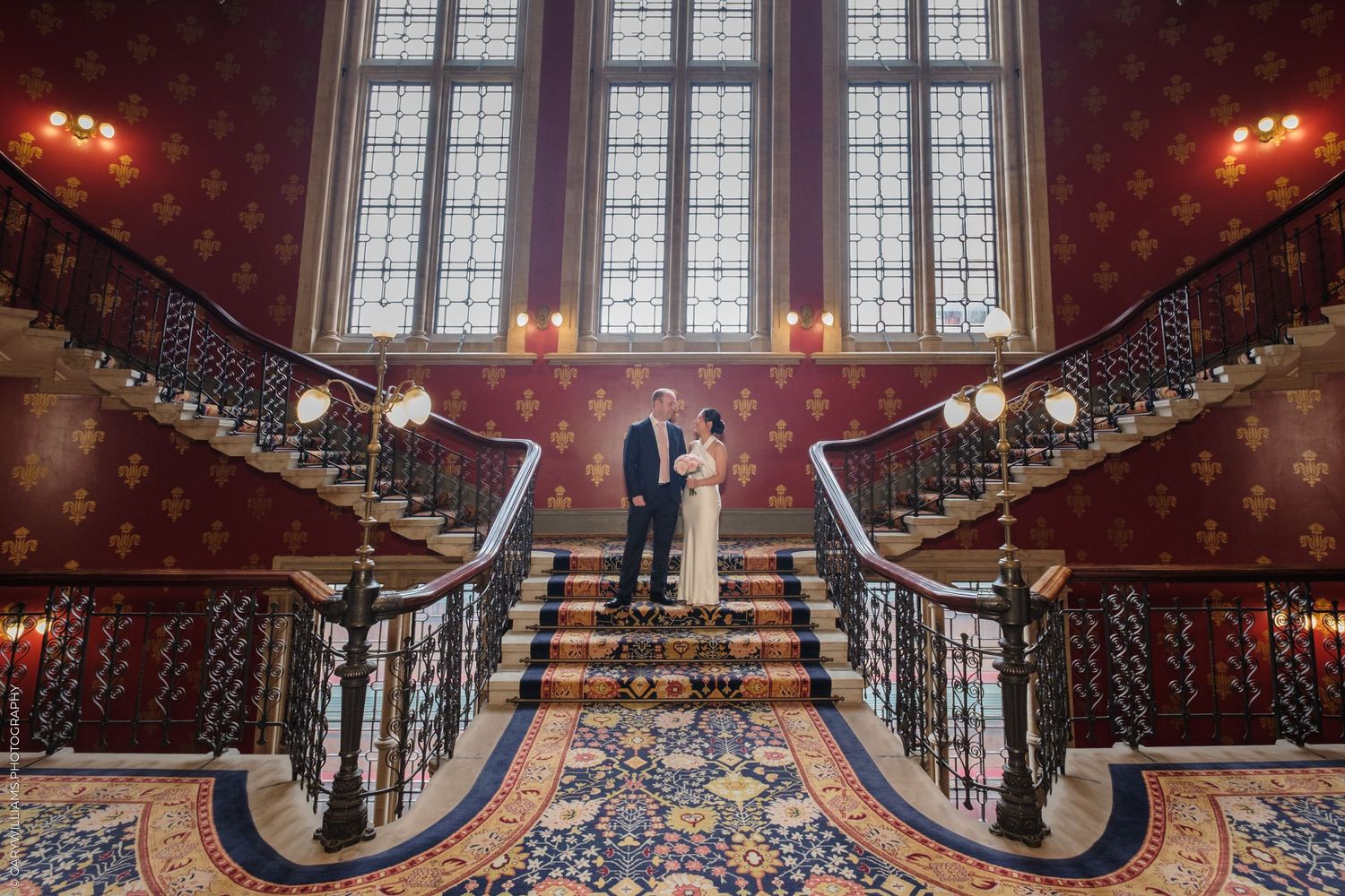 Grand staircase in historic mansion with ornate iron railings, tall windows, red walls and antique light fixtures.