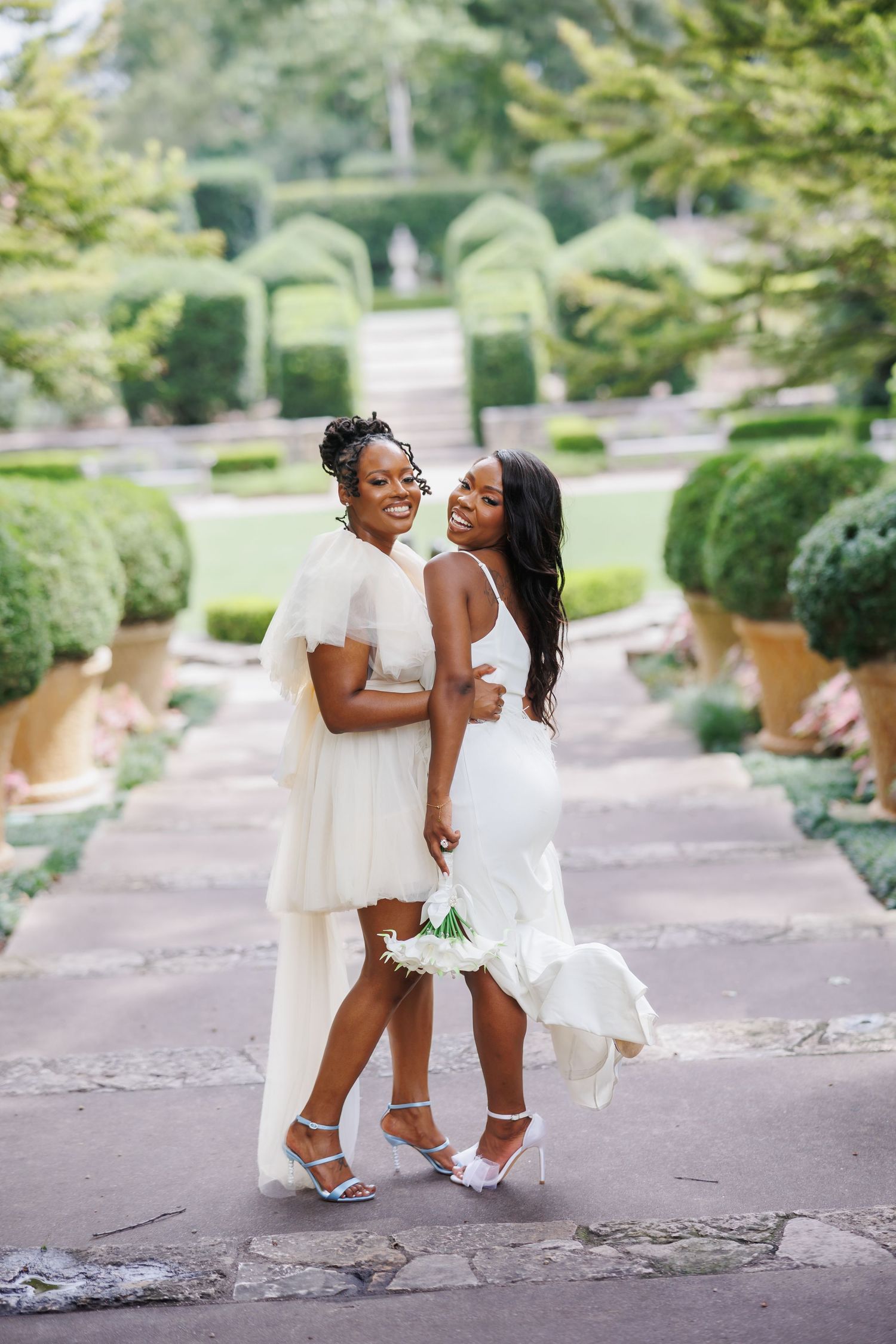 Two brides pose in front of a lush manicured garden at the Dallas Arboretum, a Dallas wedding venue