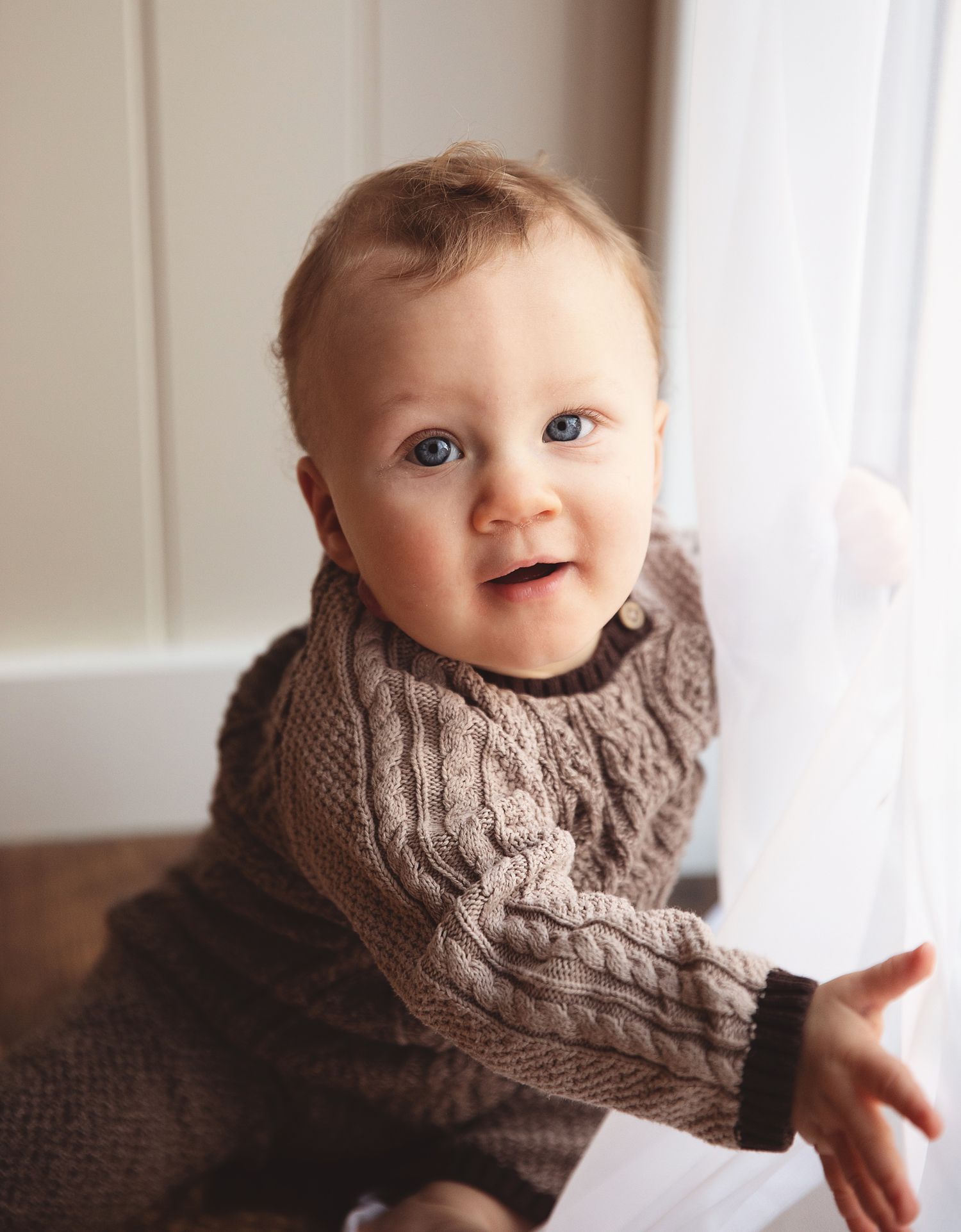 Baby wearing a cozy knit brown sweater sits near a window with a cheerful expression. During a baby photoshoot in London