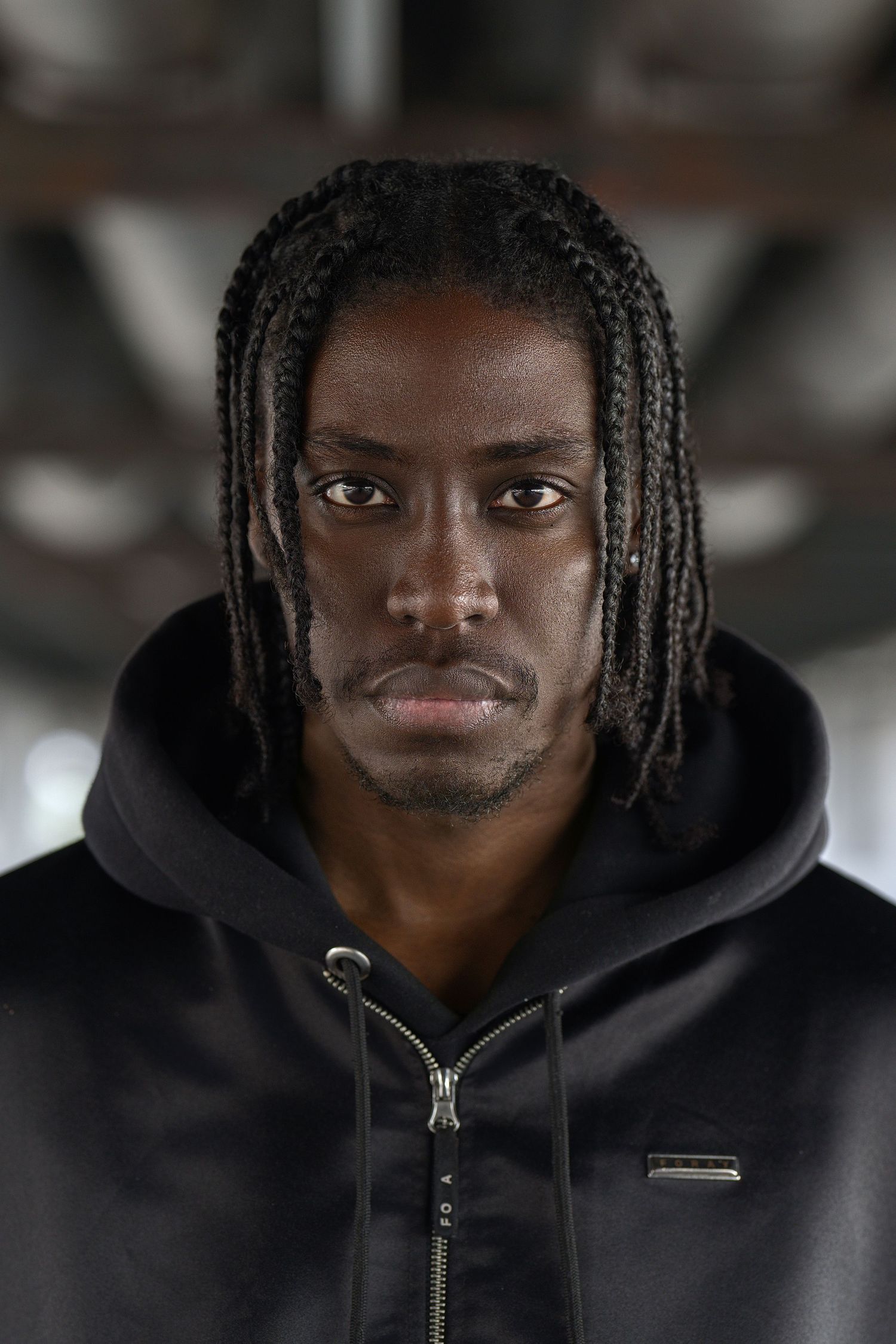 A person wearing a black hoodie with their hair in cornrow braids looks directly at the camera in a portrait shot.