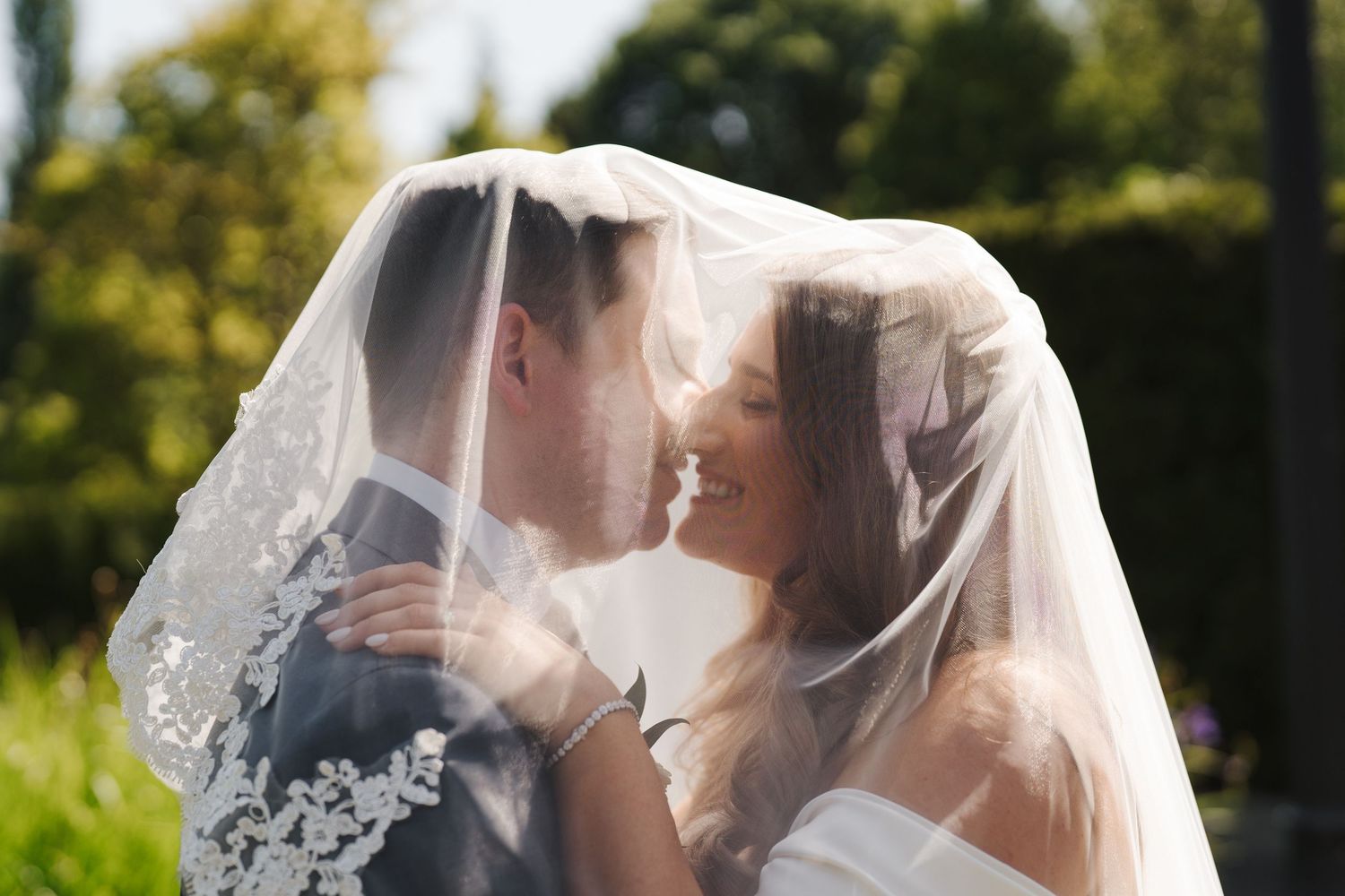An outdoor wedding ceremony taking place on a sunny day with greenery and a chapel in the background.