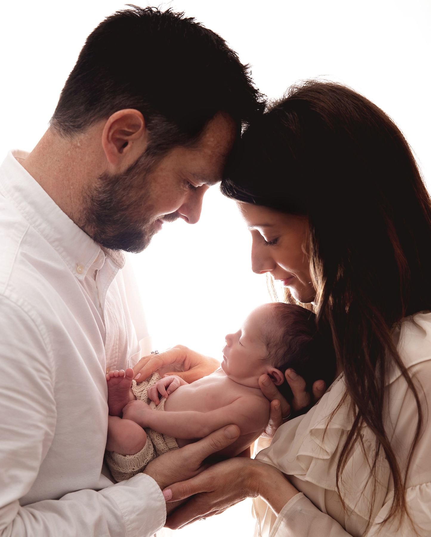 Parents lovingly holding their newborn baby against a bright backlit background in a tender family moment. At a photography studio in Bexley