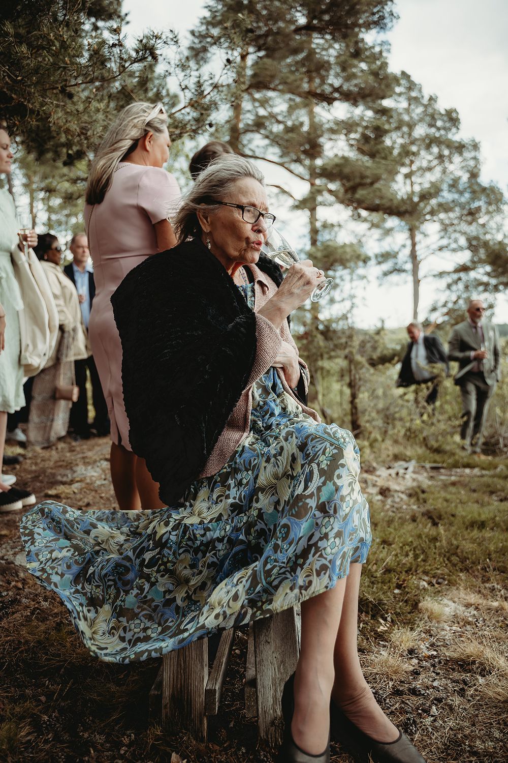 Äldre kvinna med glas champagne under bröllopsmingel vid utomhusvigsel i Sörmland – fångad av bröllopsfotograf Josefin Landhagen