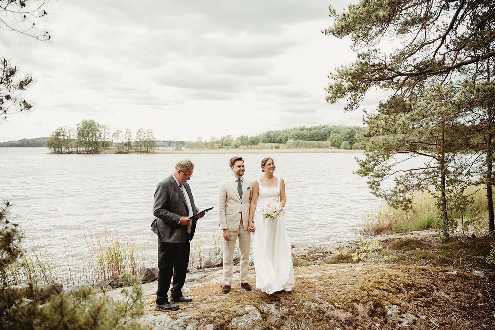 Utomhusvigsel på en klippa vid havet, brudparet står med vigselförrättare intill vattnet med skogen runt dem. Bröllopsfotografering utomhus.