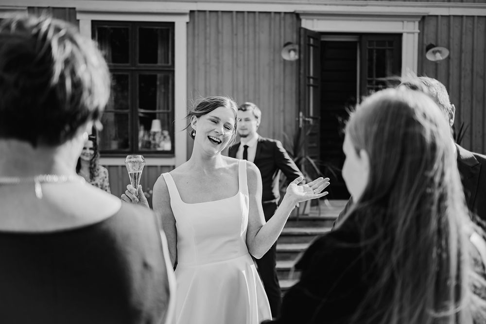 Bruden står i centrum tillsammans med gäster under mingel på bröllopsfest utomhus på en gård i Sörmland. Dokumentär porträttbild.