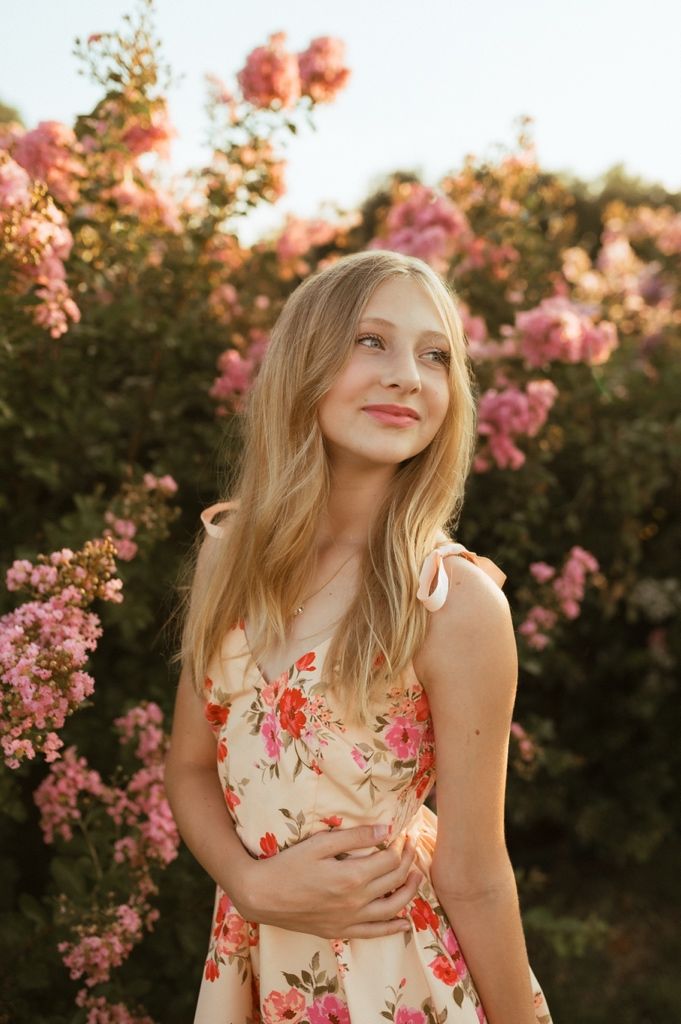 Person in floral dress poses against pink flowers at sunset in golden light.