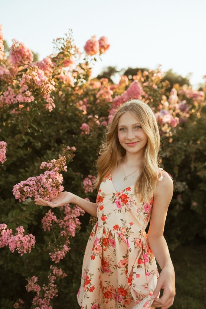 A sunset photoshoot in a blooming flower garden during golden hour captures a feminine floral dress and blooming pink blossoms.