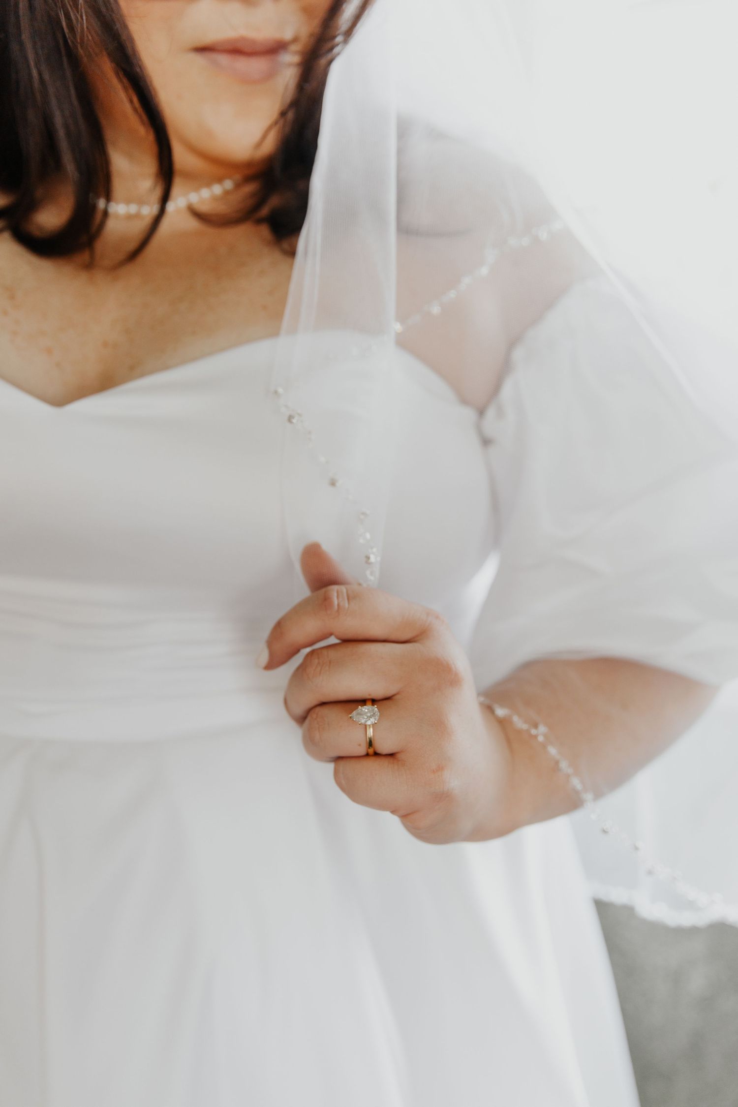 Close-up photo of hands gently holding white veil against a wedding dress with the engagement ring in focus.