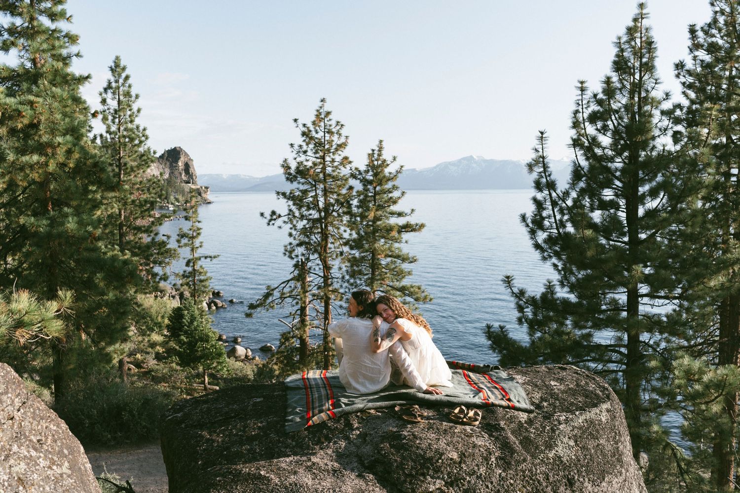 A couple sits on rocky overlook surrounded by pine trees with Lake Tahoe's blue waters and mountains in the distance.