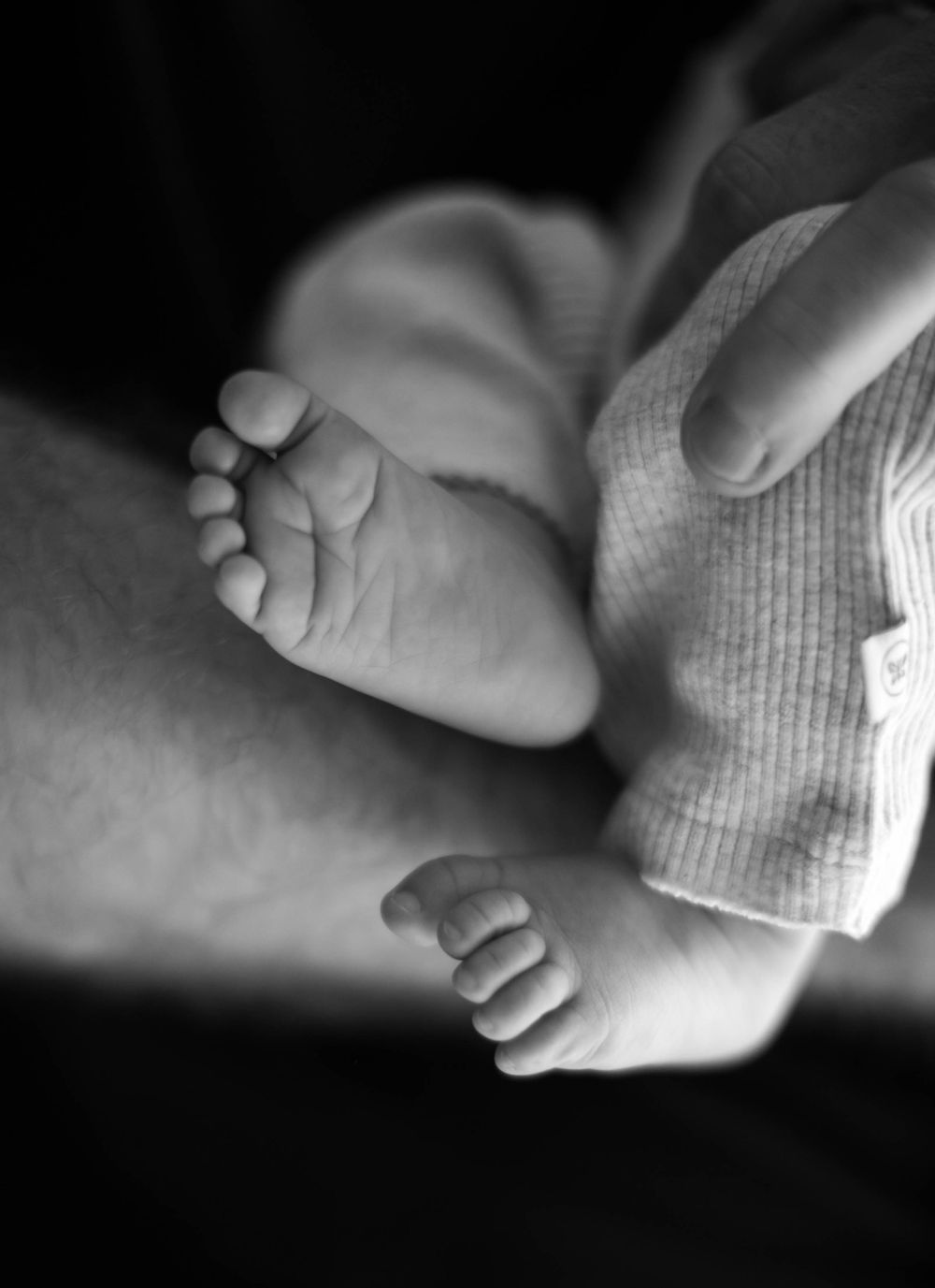 Black and white close-up photograph of tiny newborn baby feet next to fingers in gentle lighting.