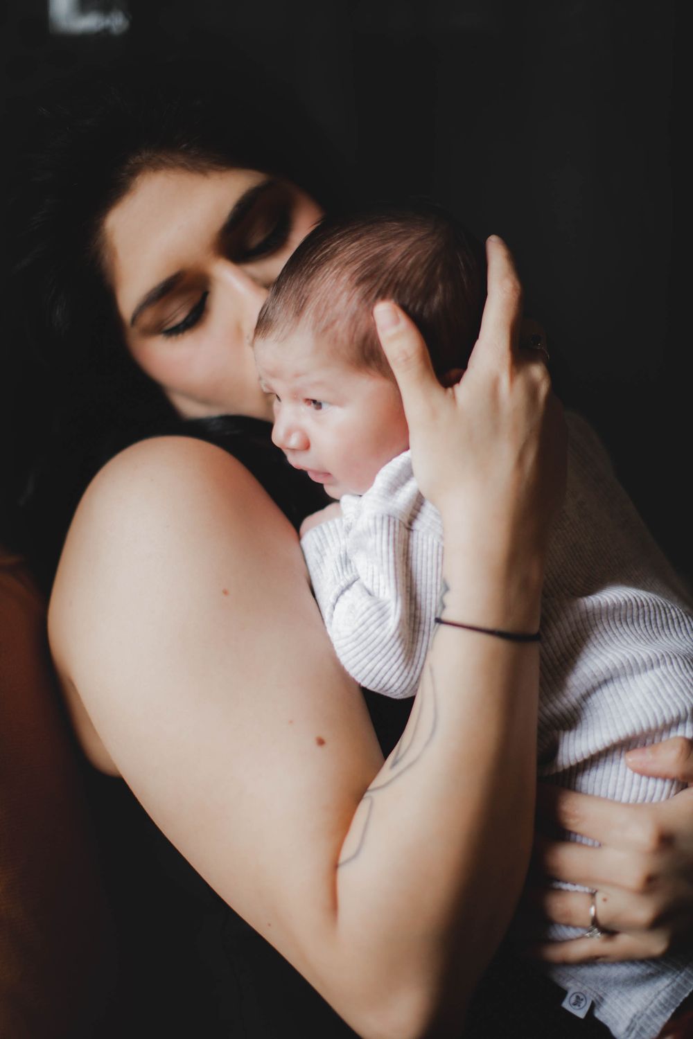 Intimate moody portrait shows tender moment between parent and baby in white knit sweater against dark background.