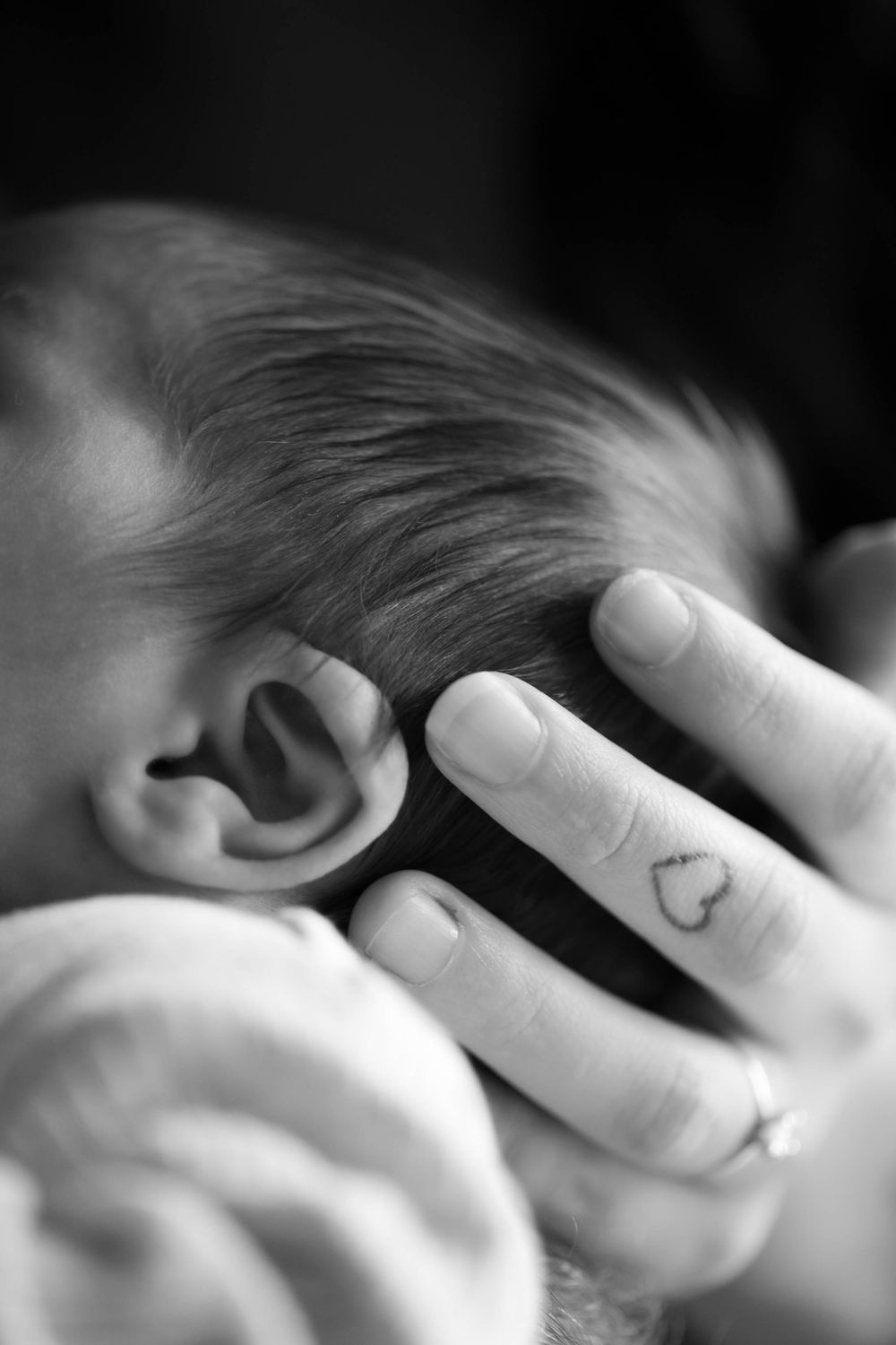 Close-up black and white photo of a hand gently touching a small baby's head, with a heart tattoo visible on the finger.