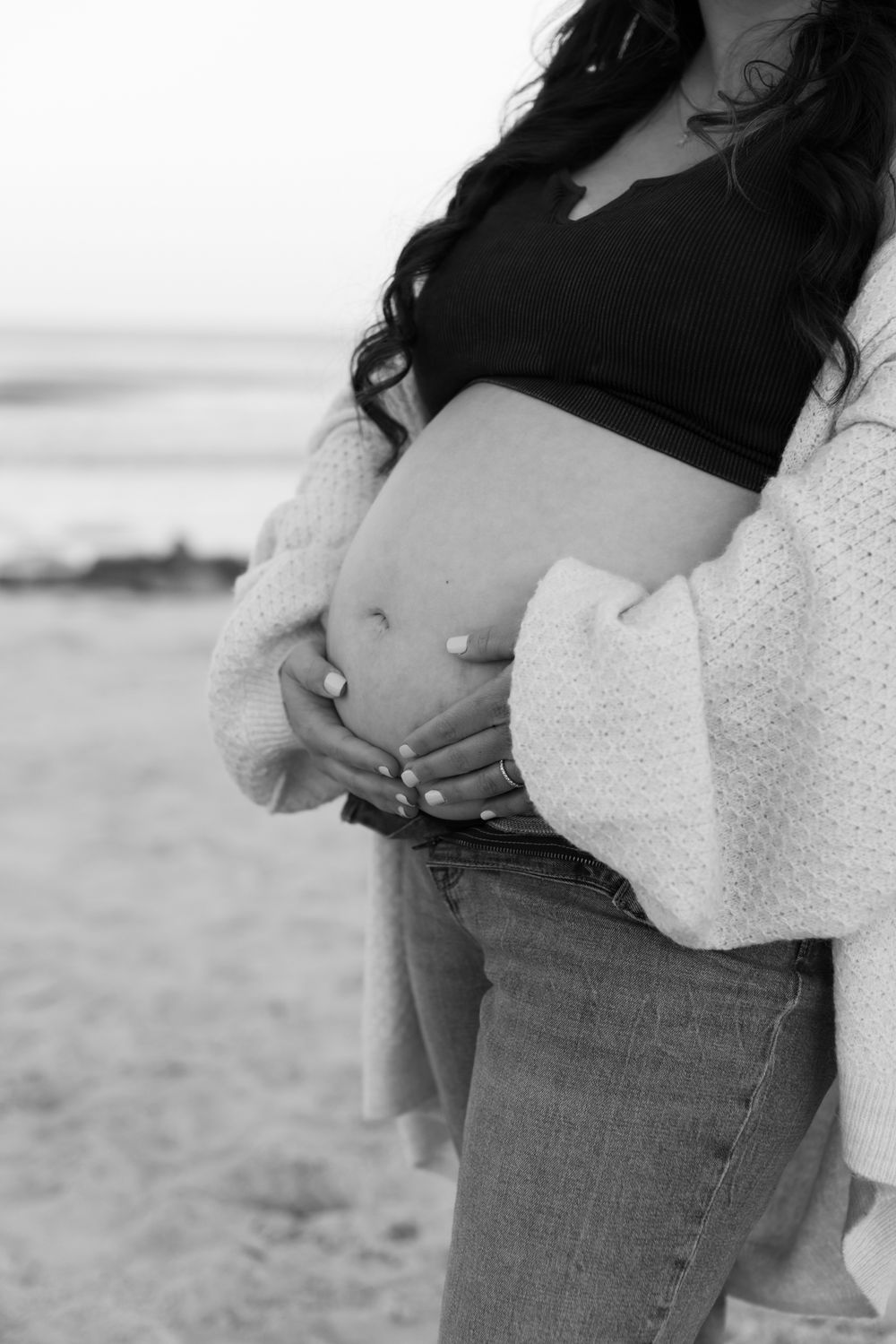 Black and white maternity photo showing pregnant belly with hands cradling bump while wearing cozy sweater on beach.