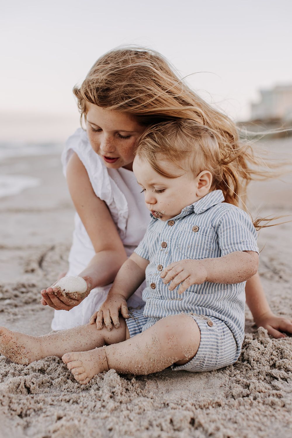 A small child wearing a blue and white striped romper plays and rolls in the sandy beach during a sunset photoshoot.