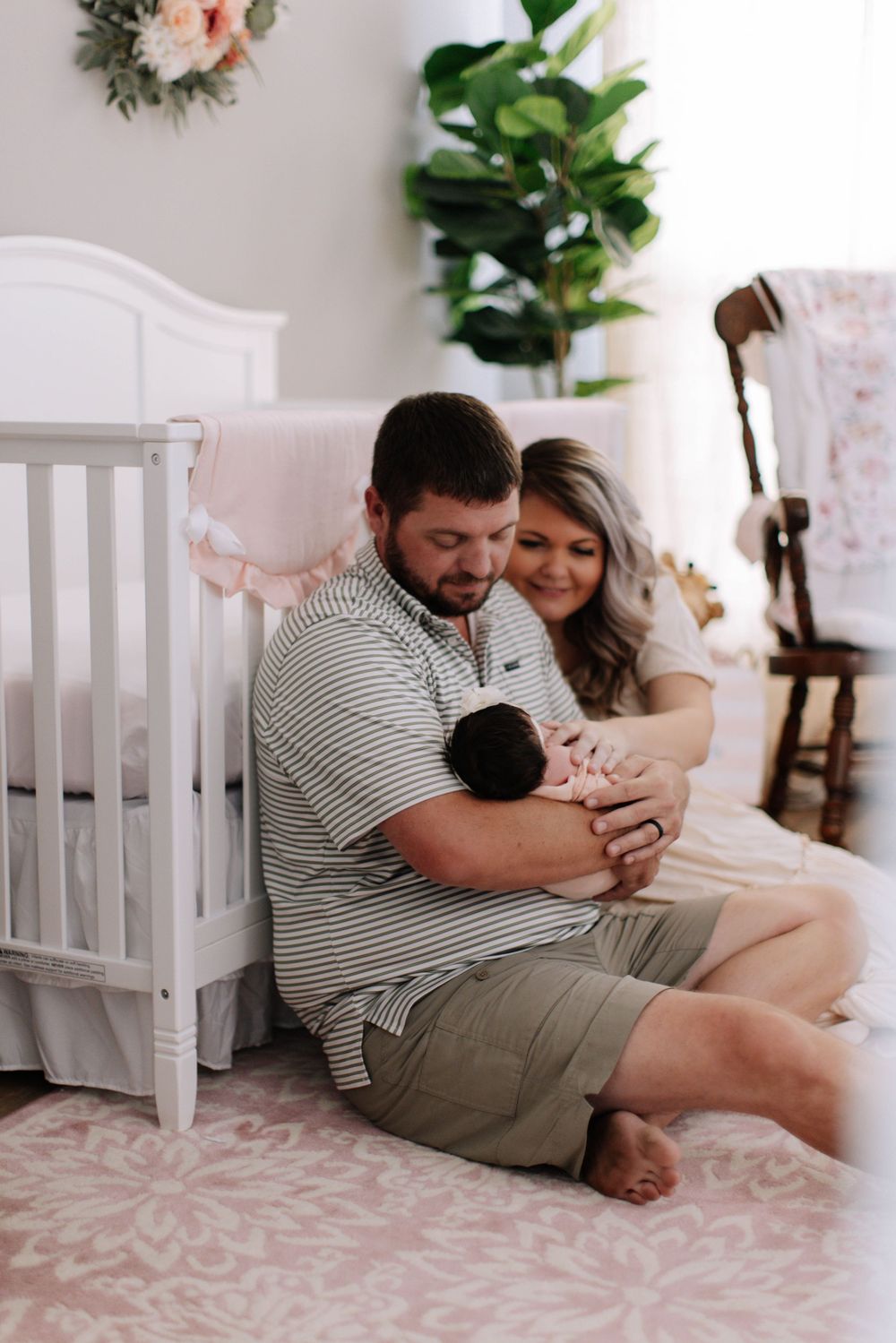 A couple sits on a pink rug next to a white crib holding a newborn baby in a cozy nursery setting.