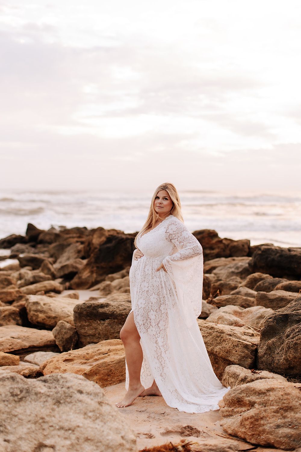 A person in a flowing white lace dress poses on rocky coastal beach during golden sunset.